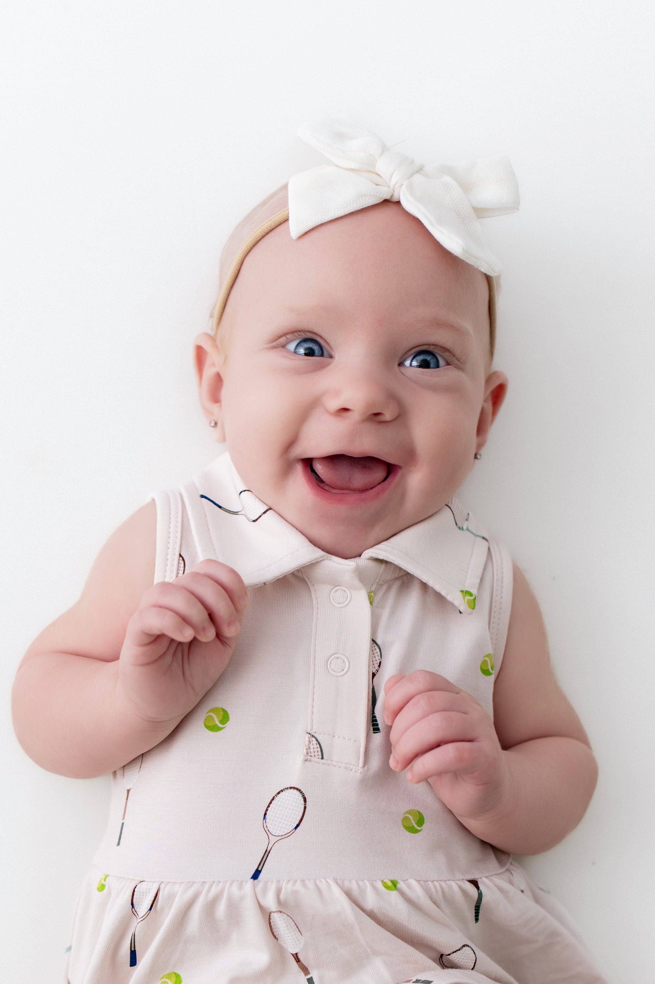 Baby wearing a white polo dress with green tennis balls and a headband on a white background