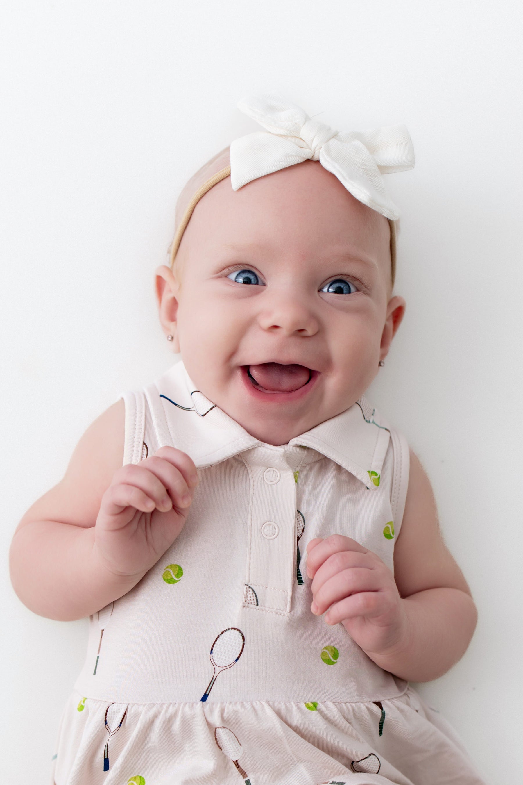Baby wearing a white polo dress with green tennis balls and a headband on a white background