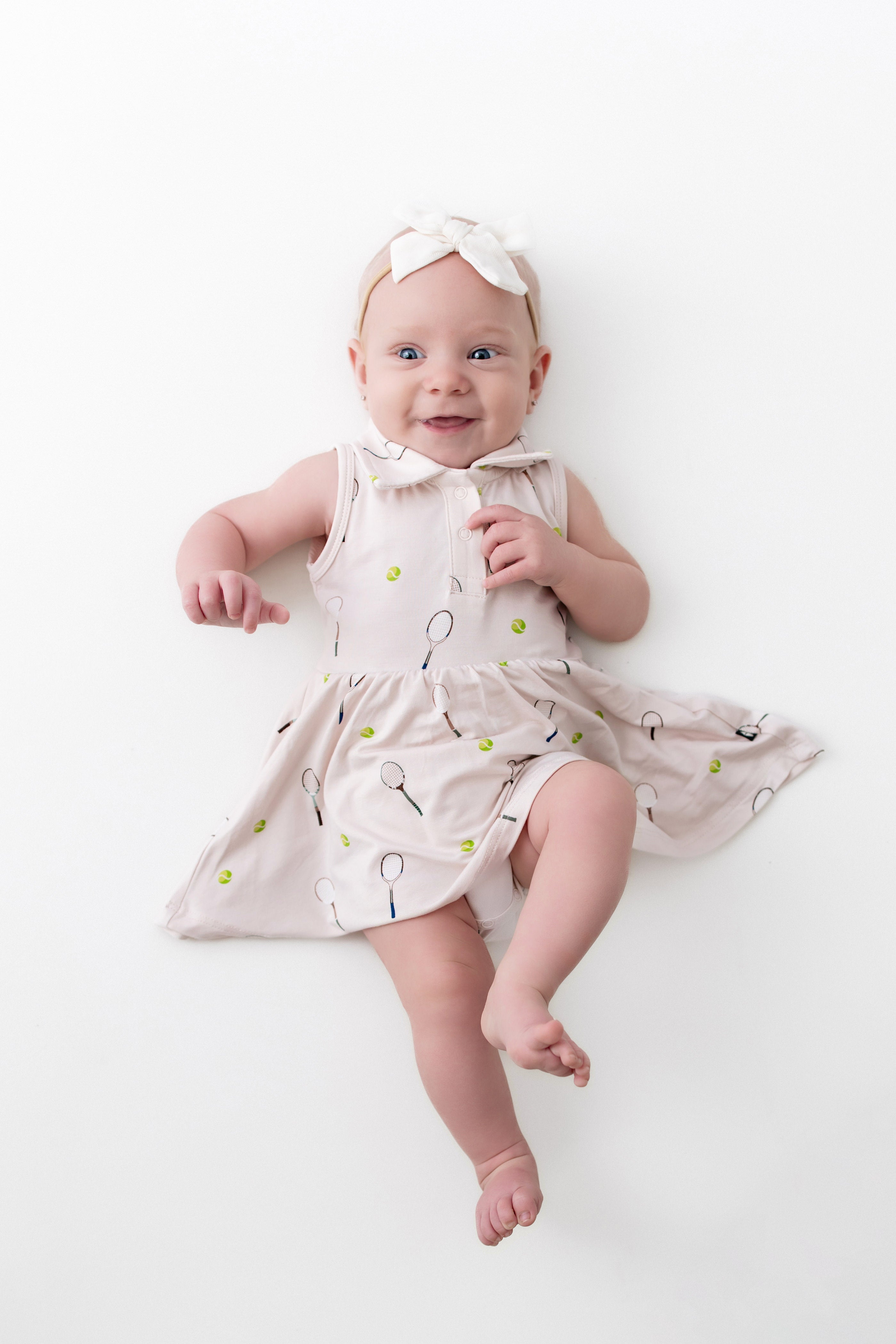 Baby wearing a light-colored dress with a tennis pattern on a white background
