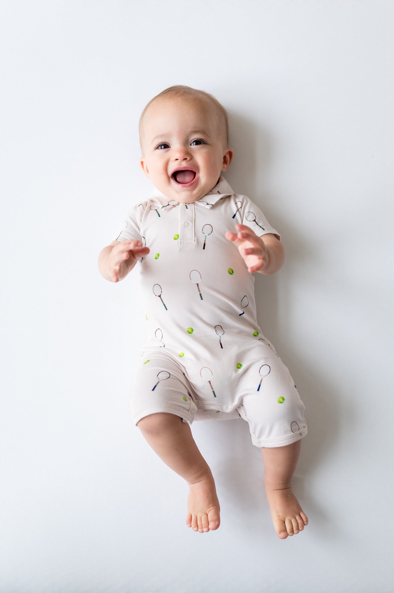 Baby wearing a white polo romper with black and green tennis patterns on a light gray background