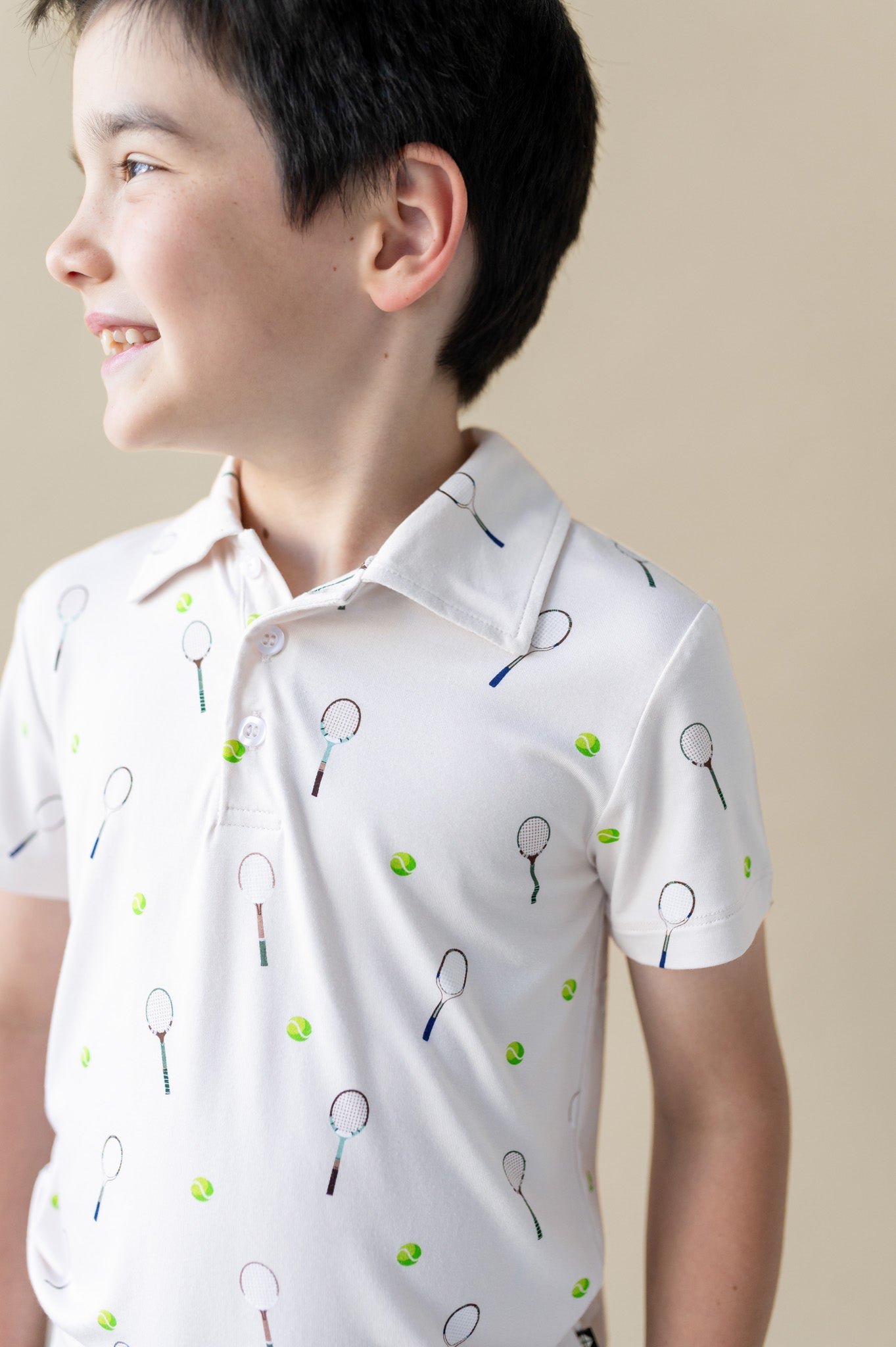 Child wearing a white polo shirt with tennis racket and ball pattern on a beige background