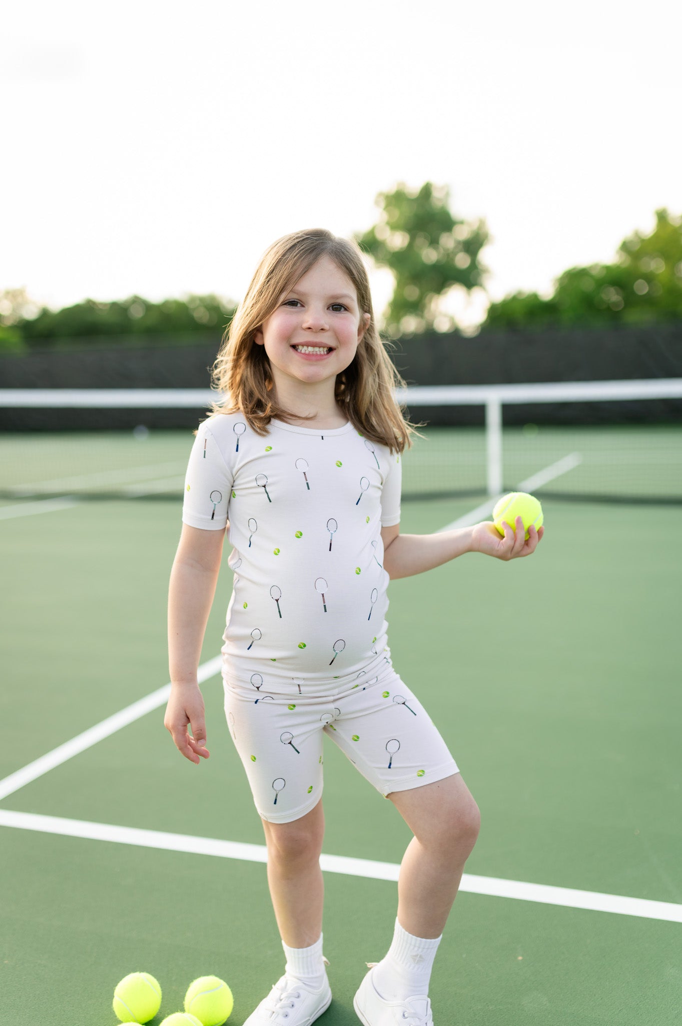 Young girl holding a tennis ball on a tennis court wearing a tennis themed short sleeve pj set