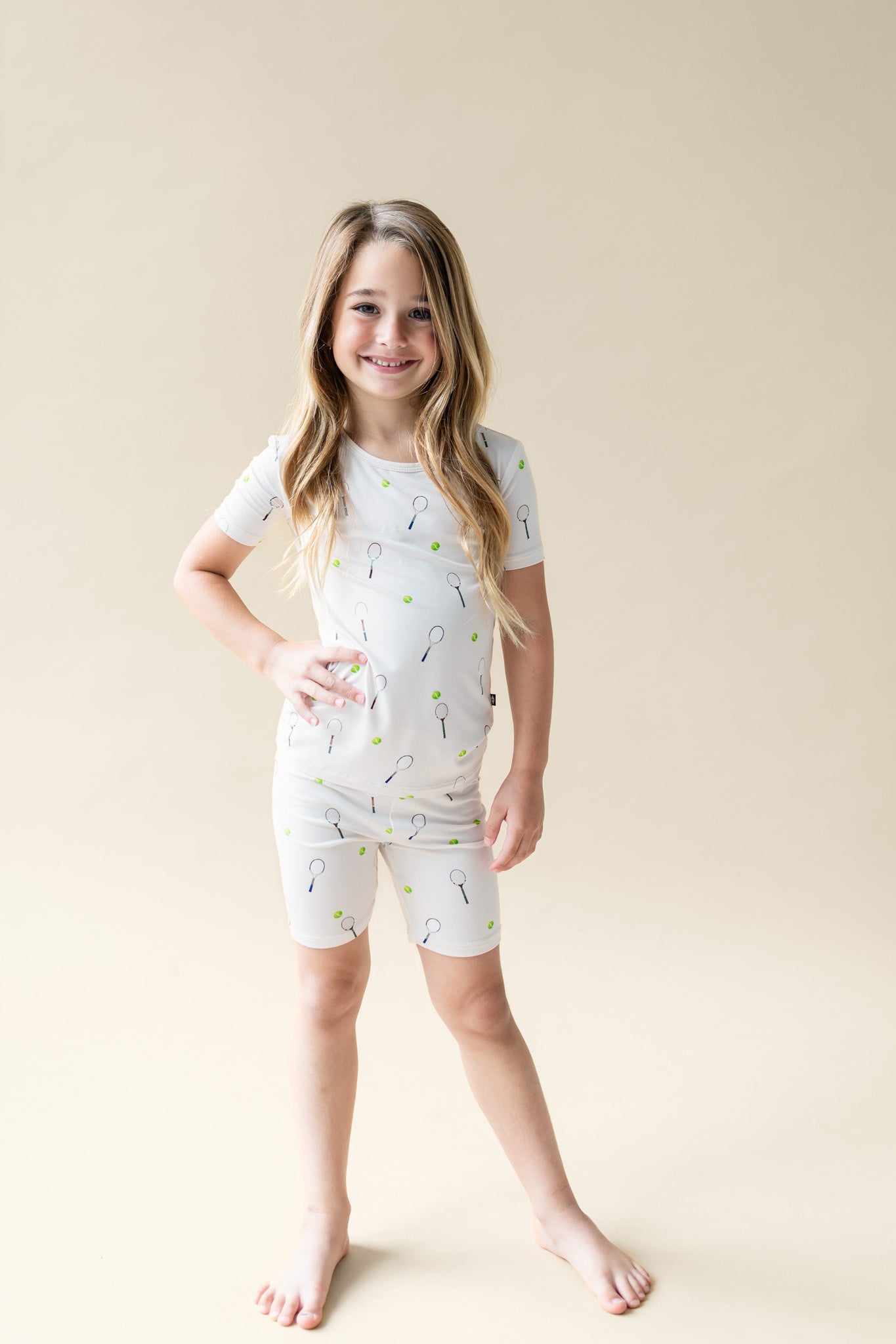 Young girl wearing a white short sleeve pajama set with a subtle tennis pattern on a beige background