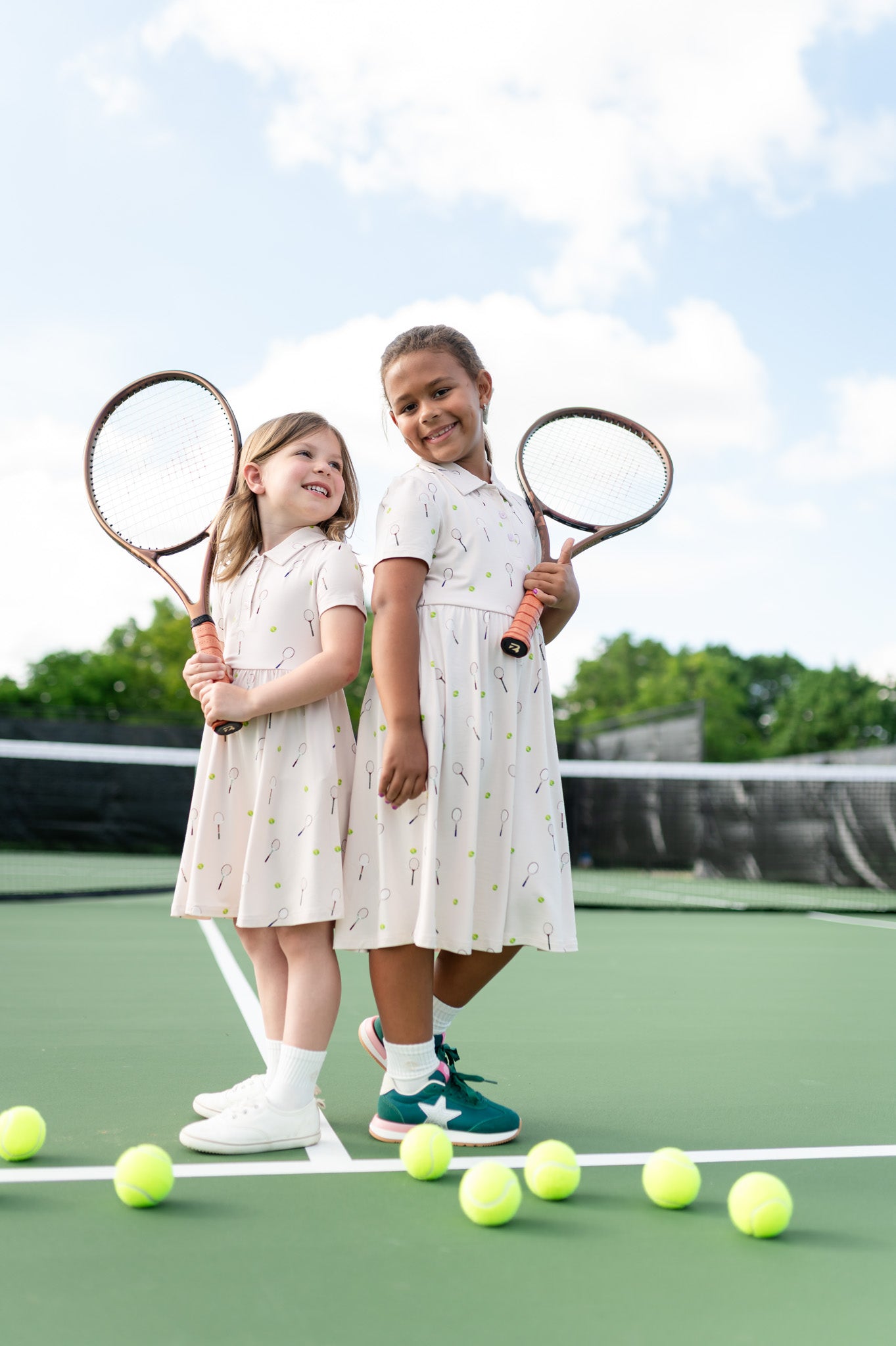 Two children on a tennis court holding tennis rackets with tennis balls around them.