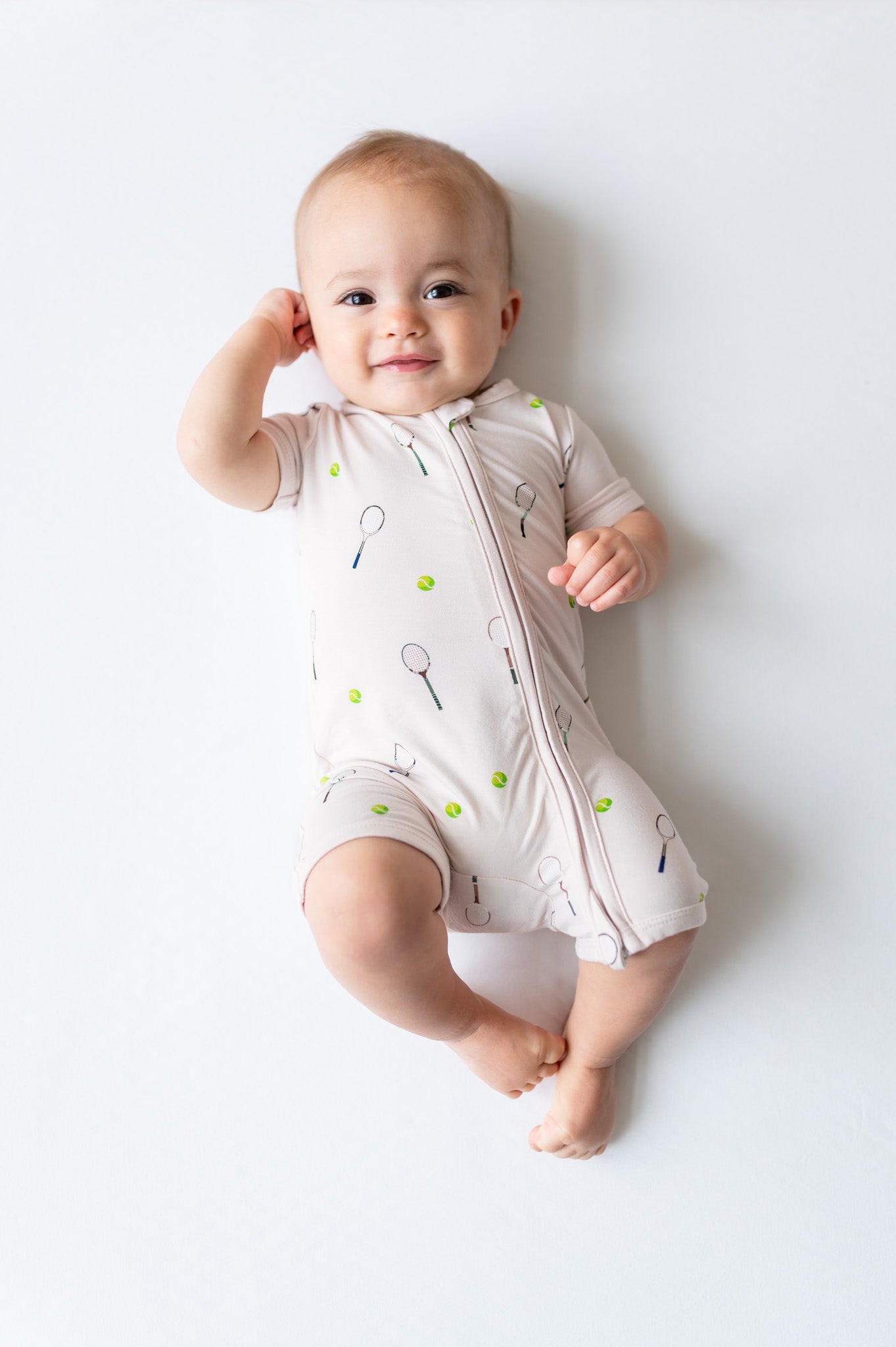 Baby wearing a white short zippered romper with tennis pattern on a light gray background