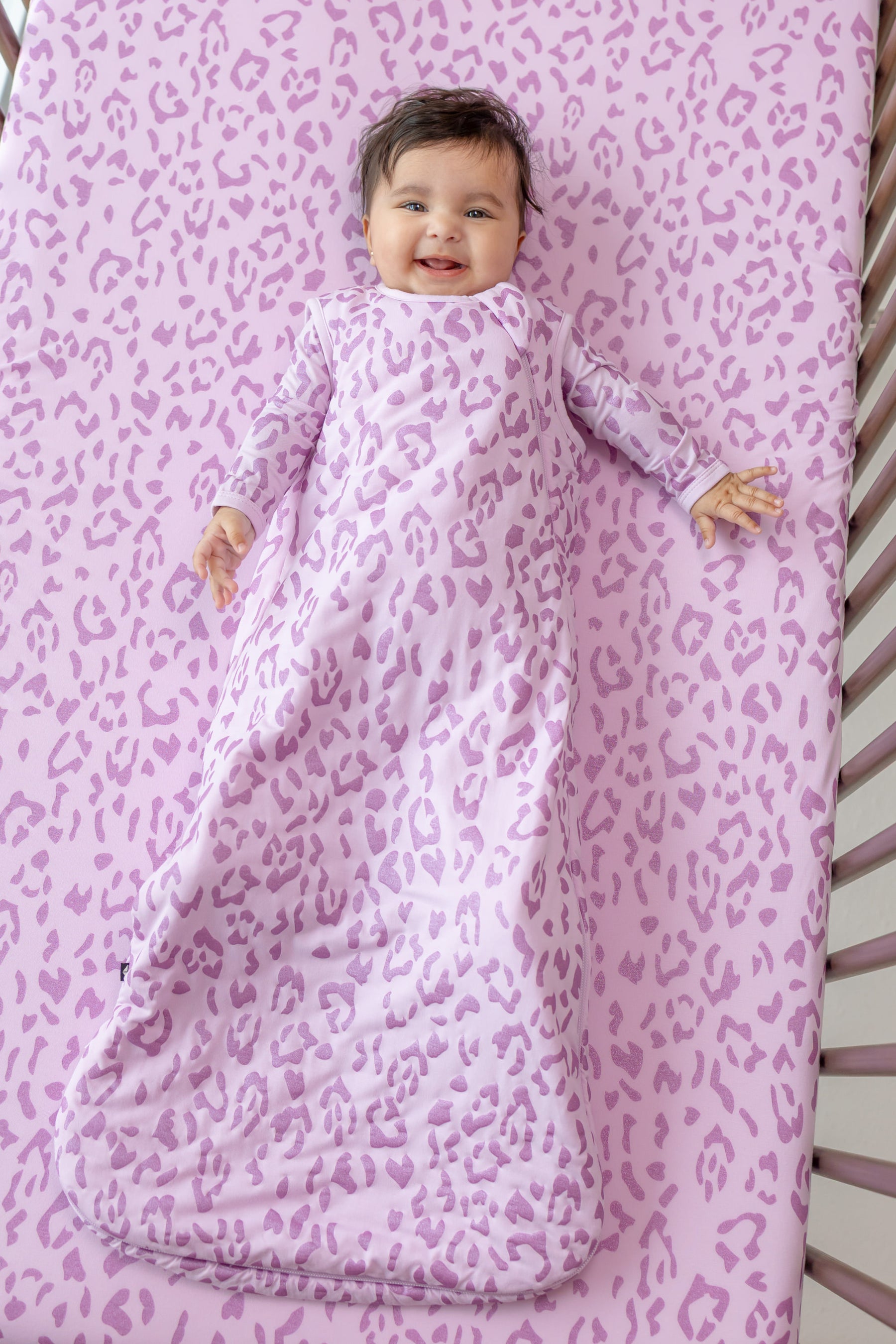 Baby wearing Thistle Leopard print sleep bag lying on a similar print crib sheet