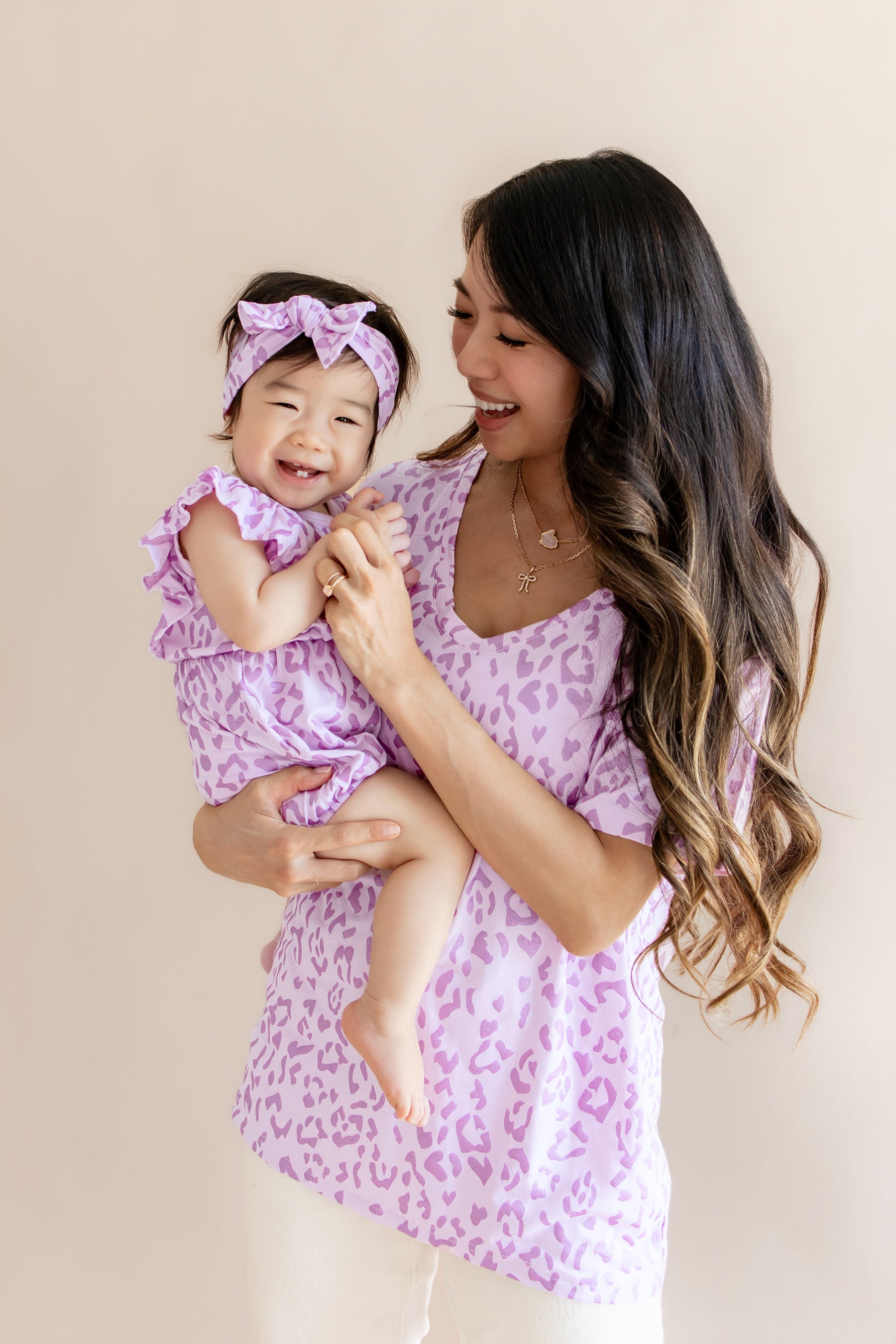 A woman holding a smiling toddler, both wearing matching light purple and pink leopard-print outfits against a soft beige studio background.