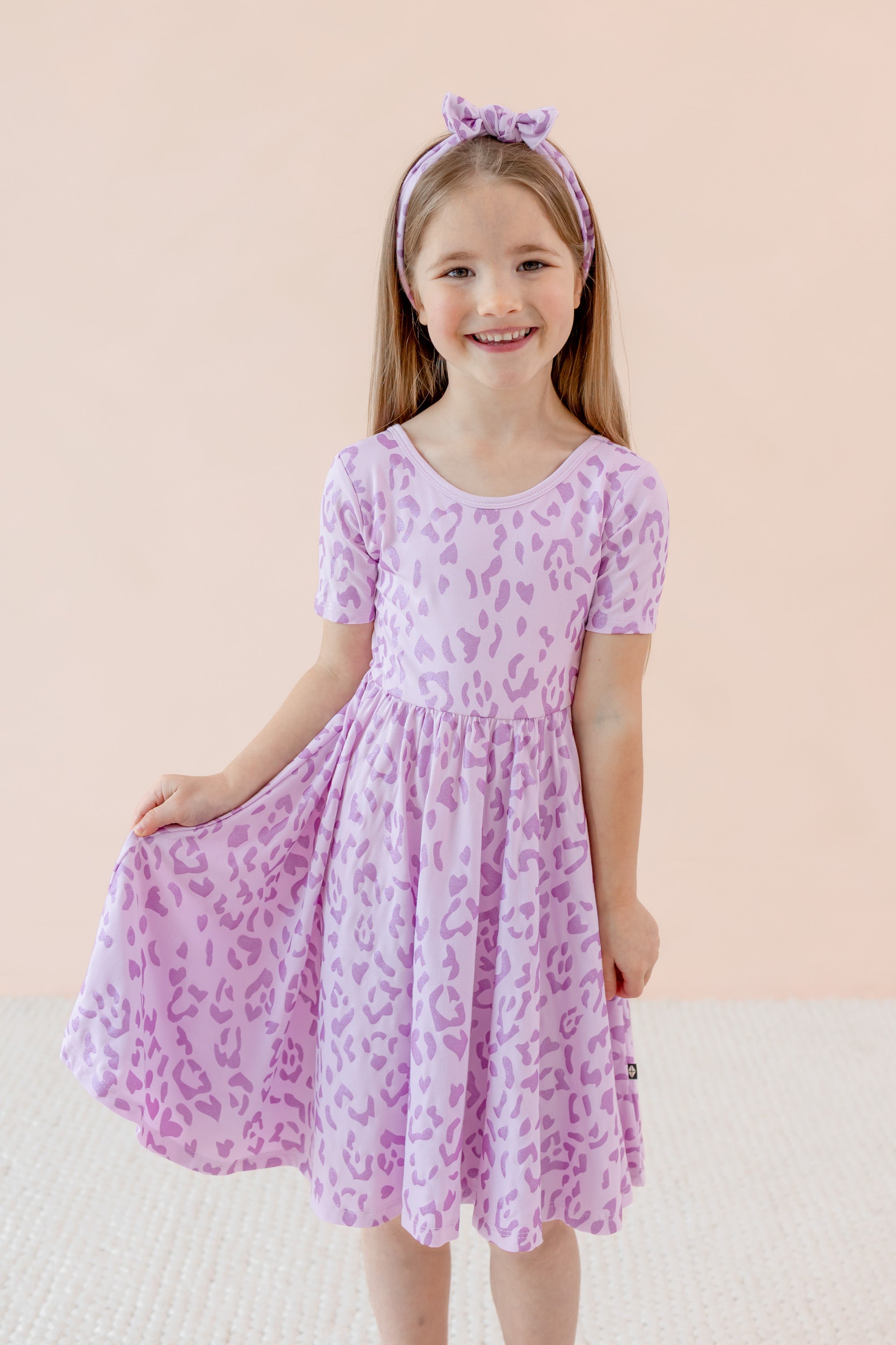 Child wearing Twirl dress in Thistle Leopard print and similar print bow headband on a beige background
