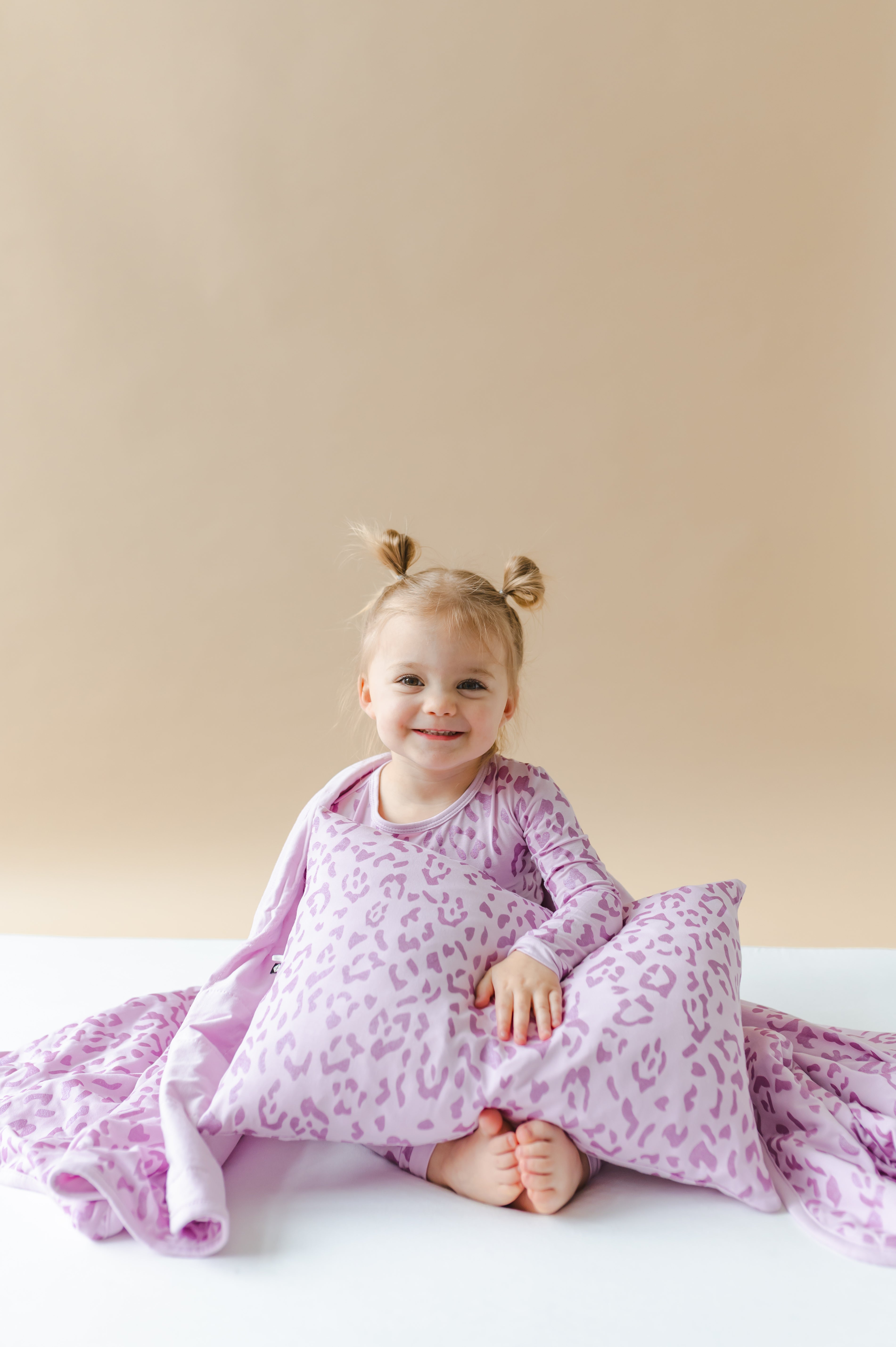 Child holding a Toddler Pillowcase in Thistle Leopard print with a toddler blanket, wearing a similar print long sleeve pajama set on a white sheet