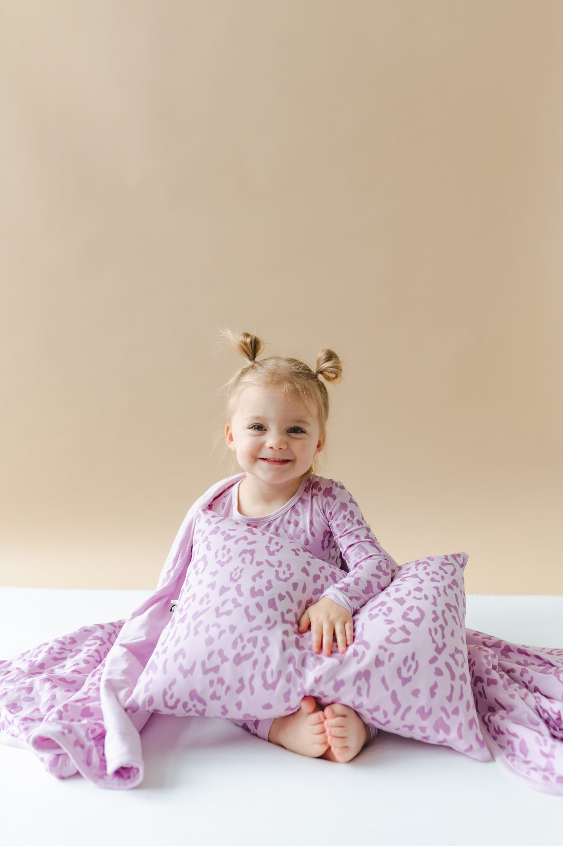 Child holding a Toddler Pillowcase in Thistle Leopard print with a toddler blanket, wearing a similar print long sleeve pajama set on a white sheet