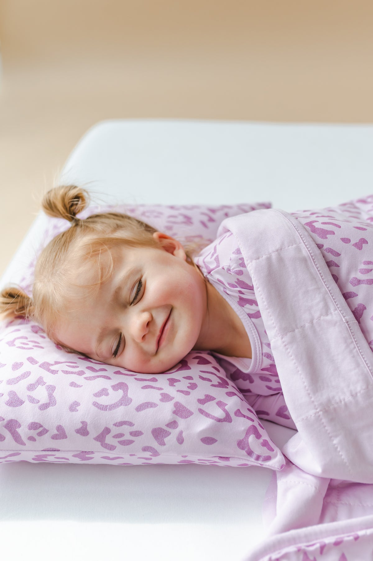 Child lying on a Thistle Leopard print toddler pillowcase wearing a similar print shirt under a thistle print Toddler blanket on a white sheet