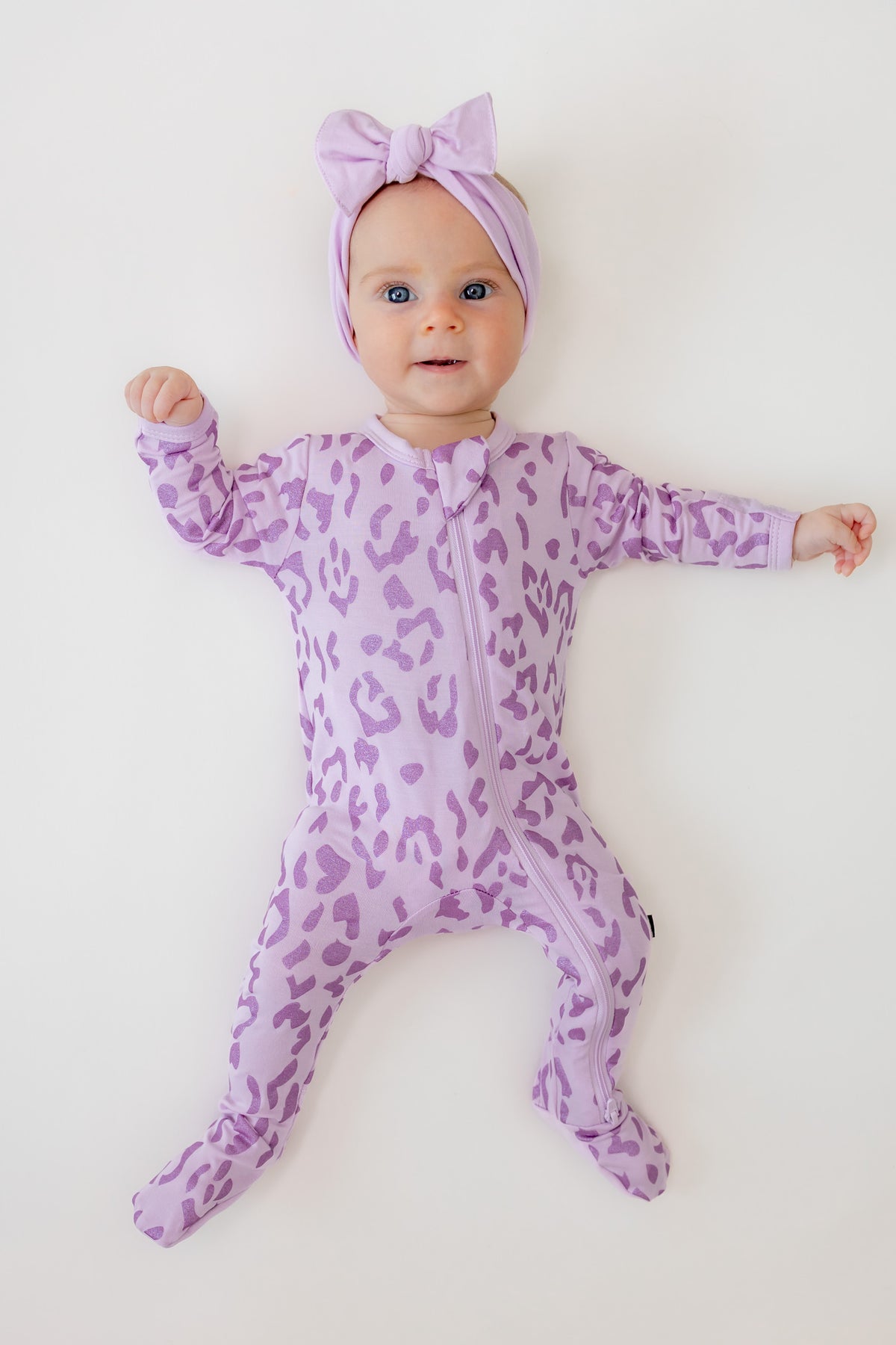 Baby wearing zippered footie in Thistle Leopard print with a thistle bow headband on a beige background