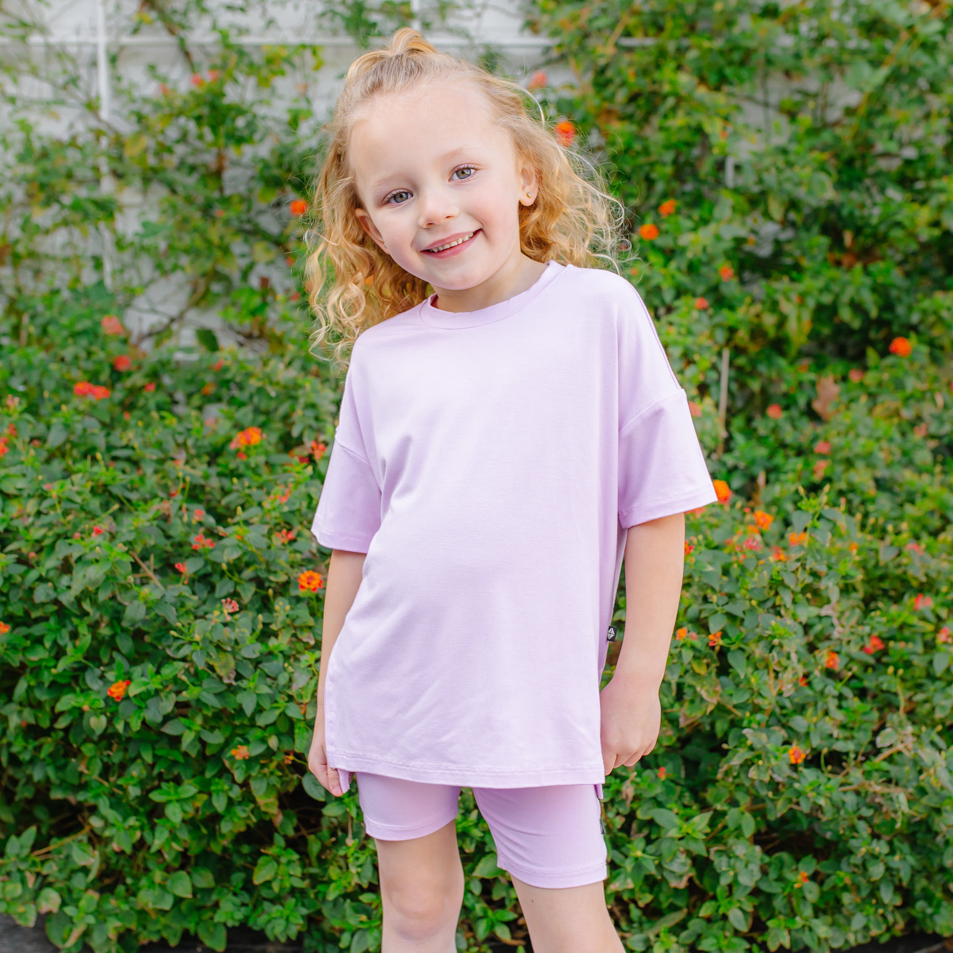 Young girl in a purple biker short set standing in front of green foliage with orange flowers.