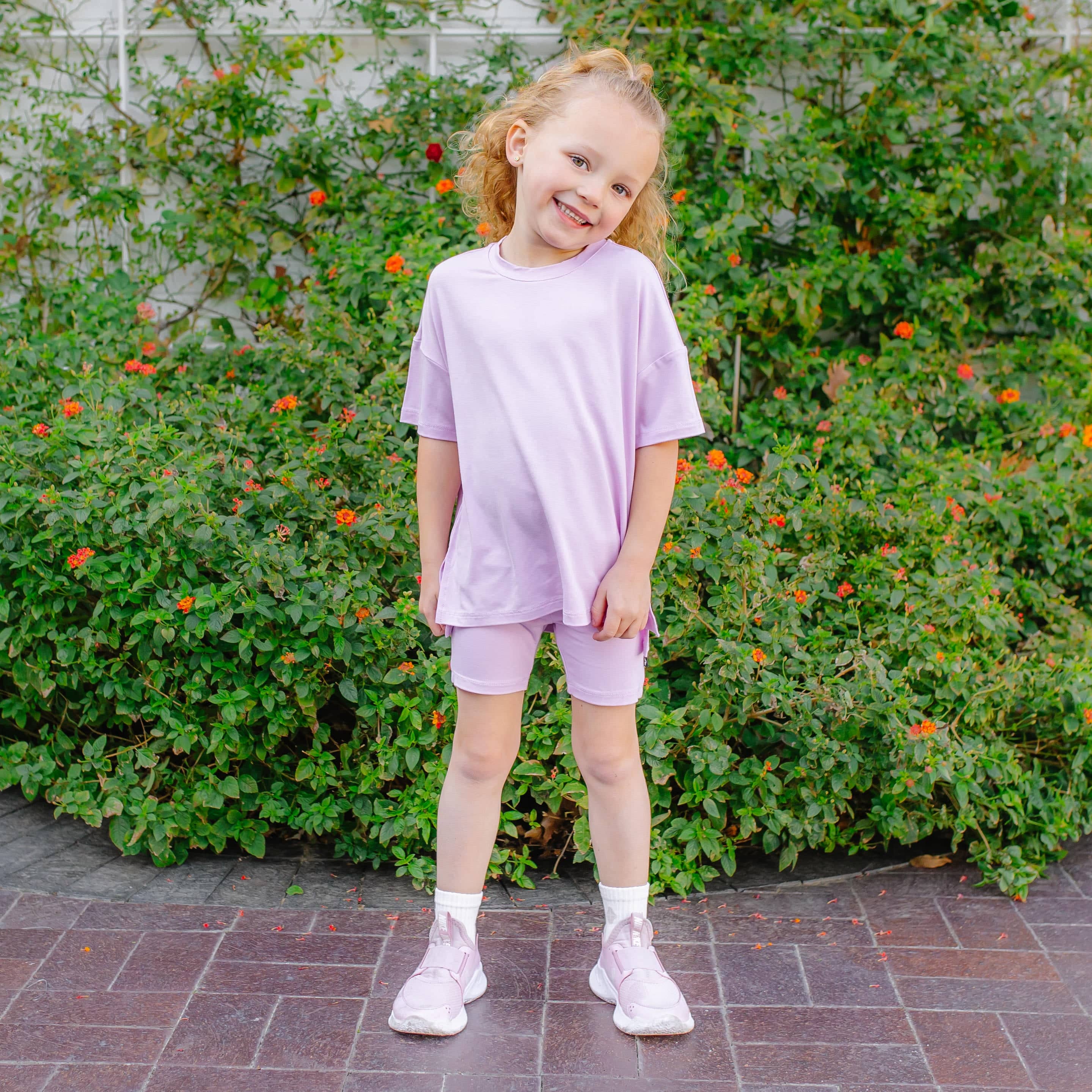 Young girl in a lavender outfit standing in front of green bushes with orange flowers.