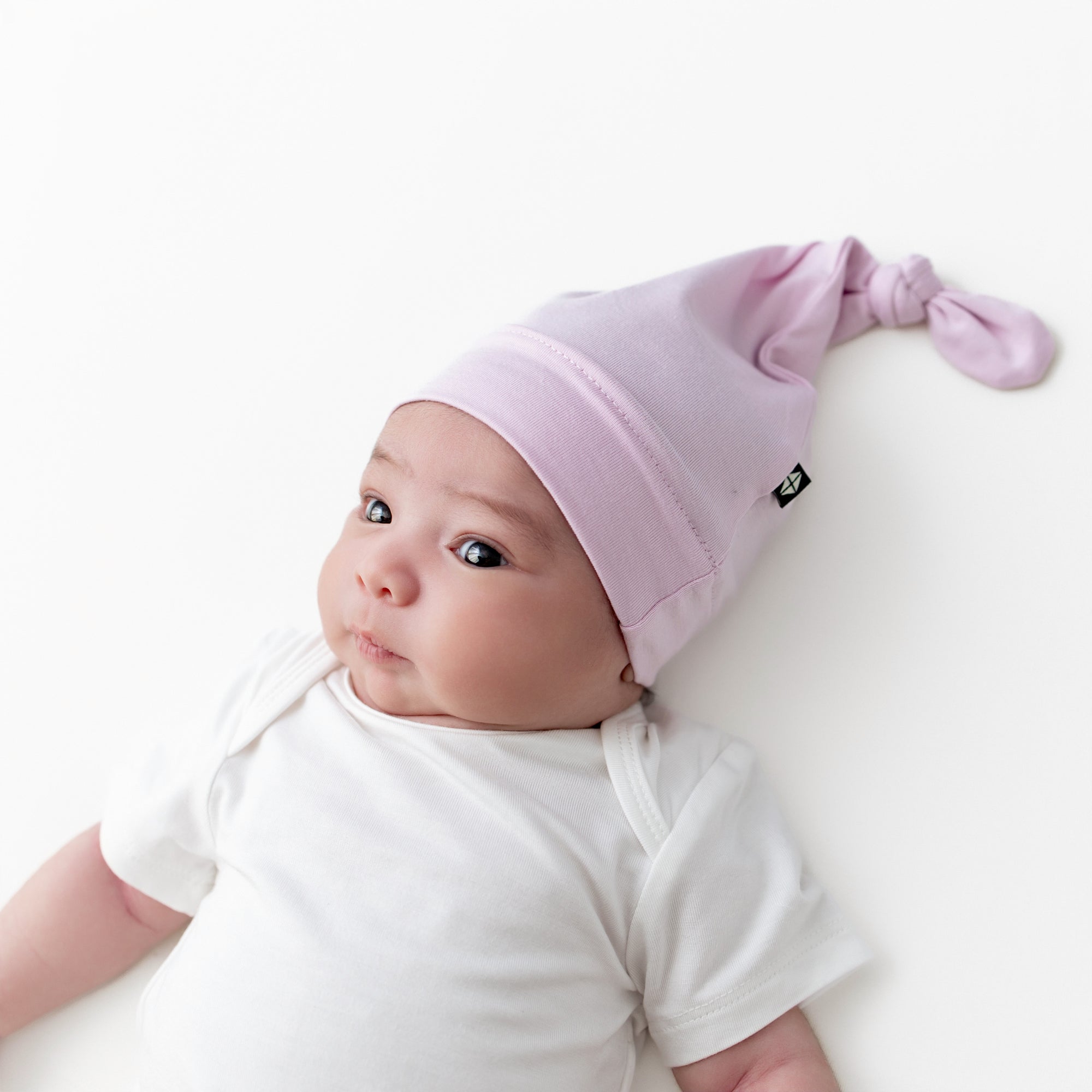 Infant girl wearing the Knotted Cap in Thistle with a white bodysuit laying on a white surface
