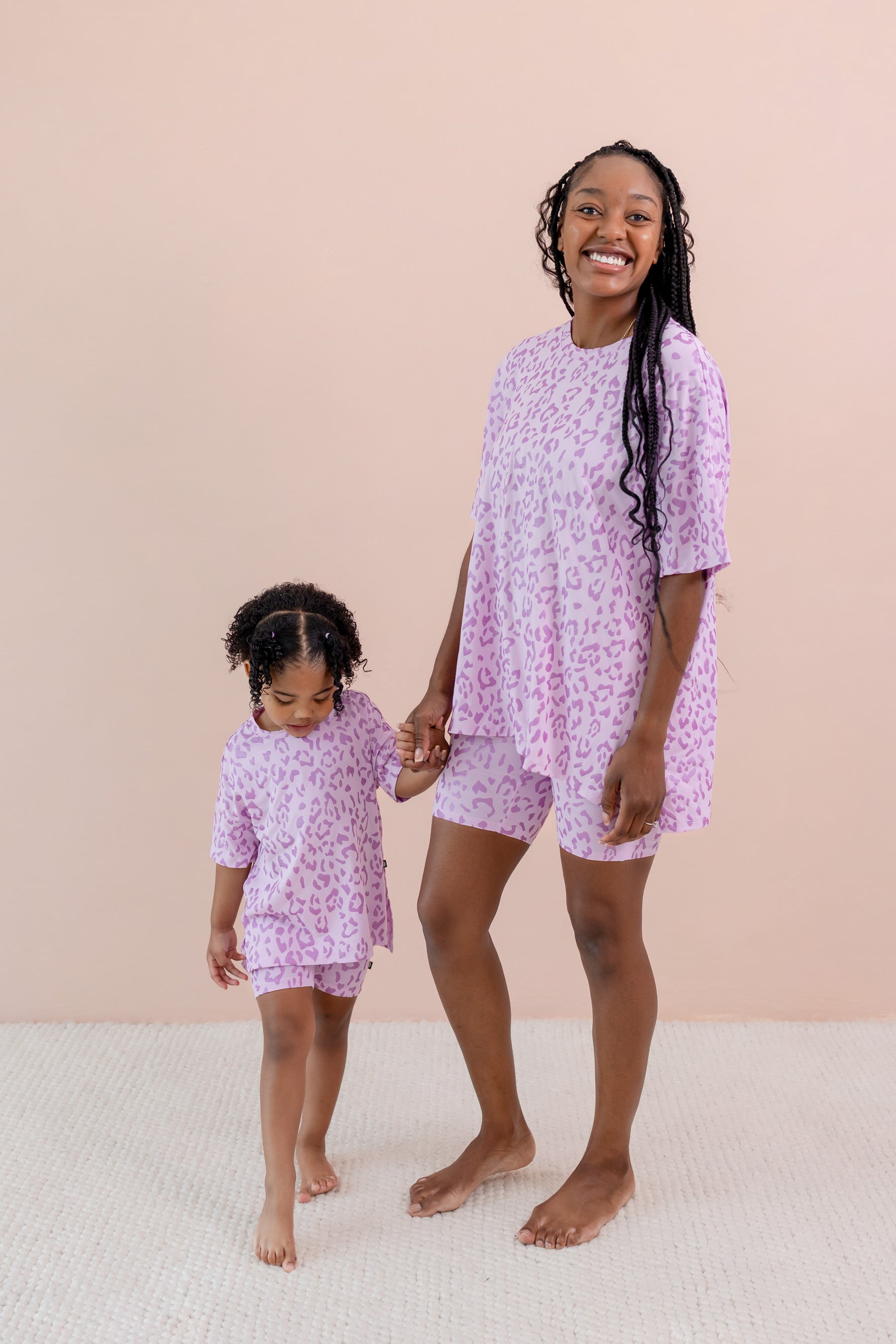 A woman and a young girl wearing matching light purple short-sleeve biker sets with a small pink animal print, standing barefoot against a solid pale peach background.