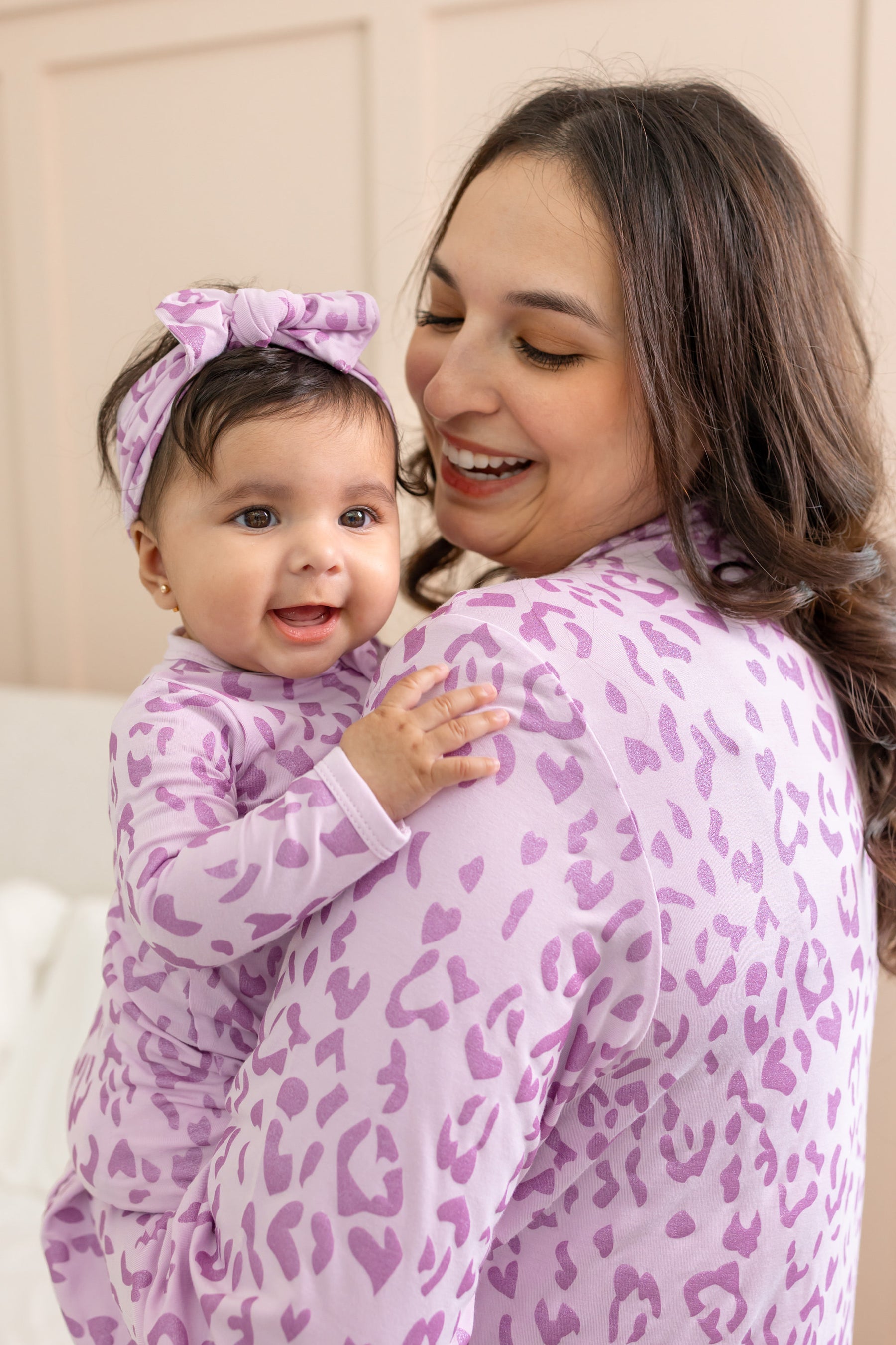 A smiling woman and baby wearing matching light purple and pink animal-print outfits, with the baby also wearing a matching bow headband in a cozy indoor setting.