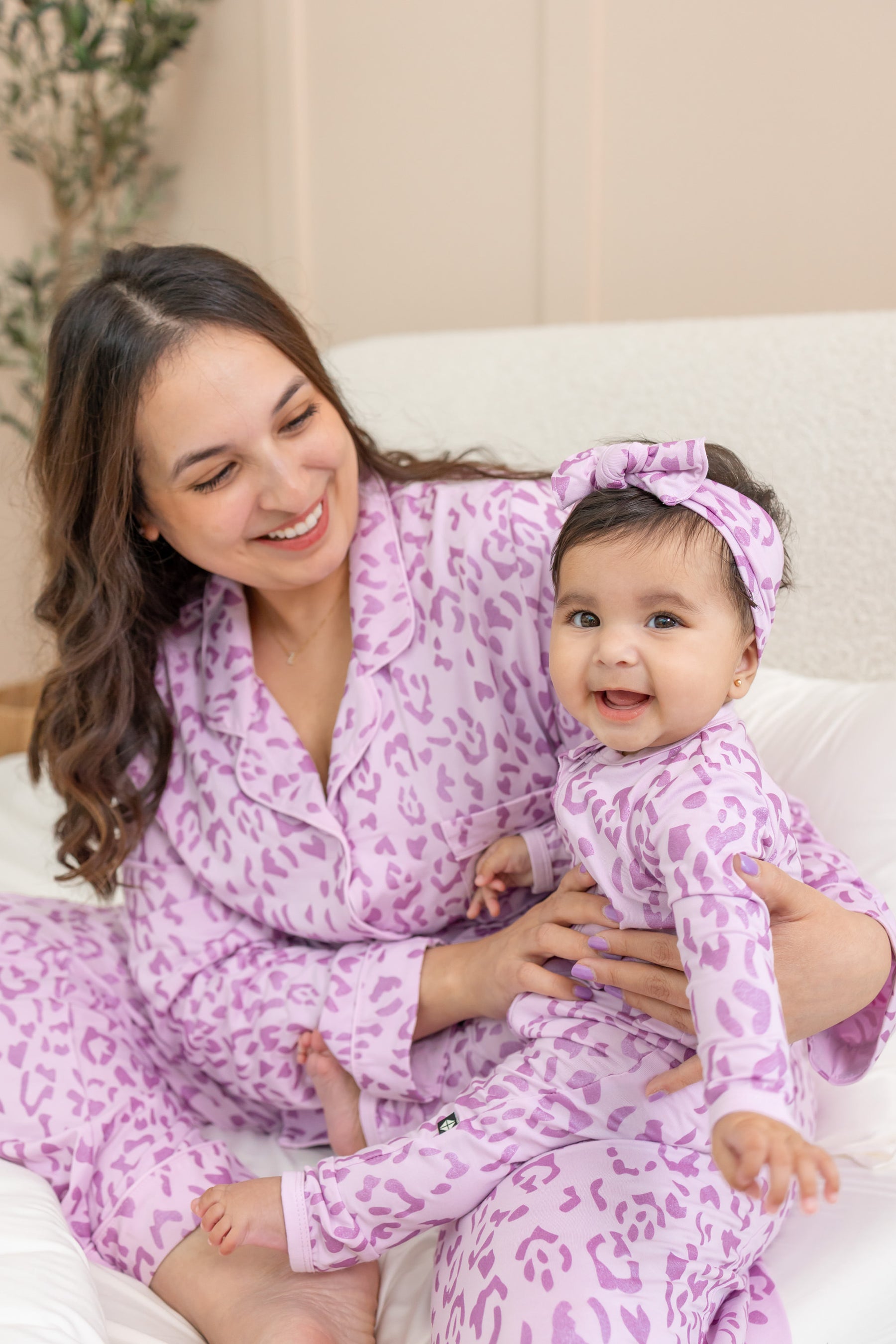 A smiling woman and baby wearing matching light purple and pink leopard-print outfits and headband set, sitting together on a white sofa.