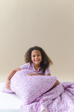 Child holding a Thistle Leopard print Standard pillowcase and blanket wearing a similar print shirt on a white sheet