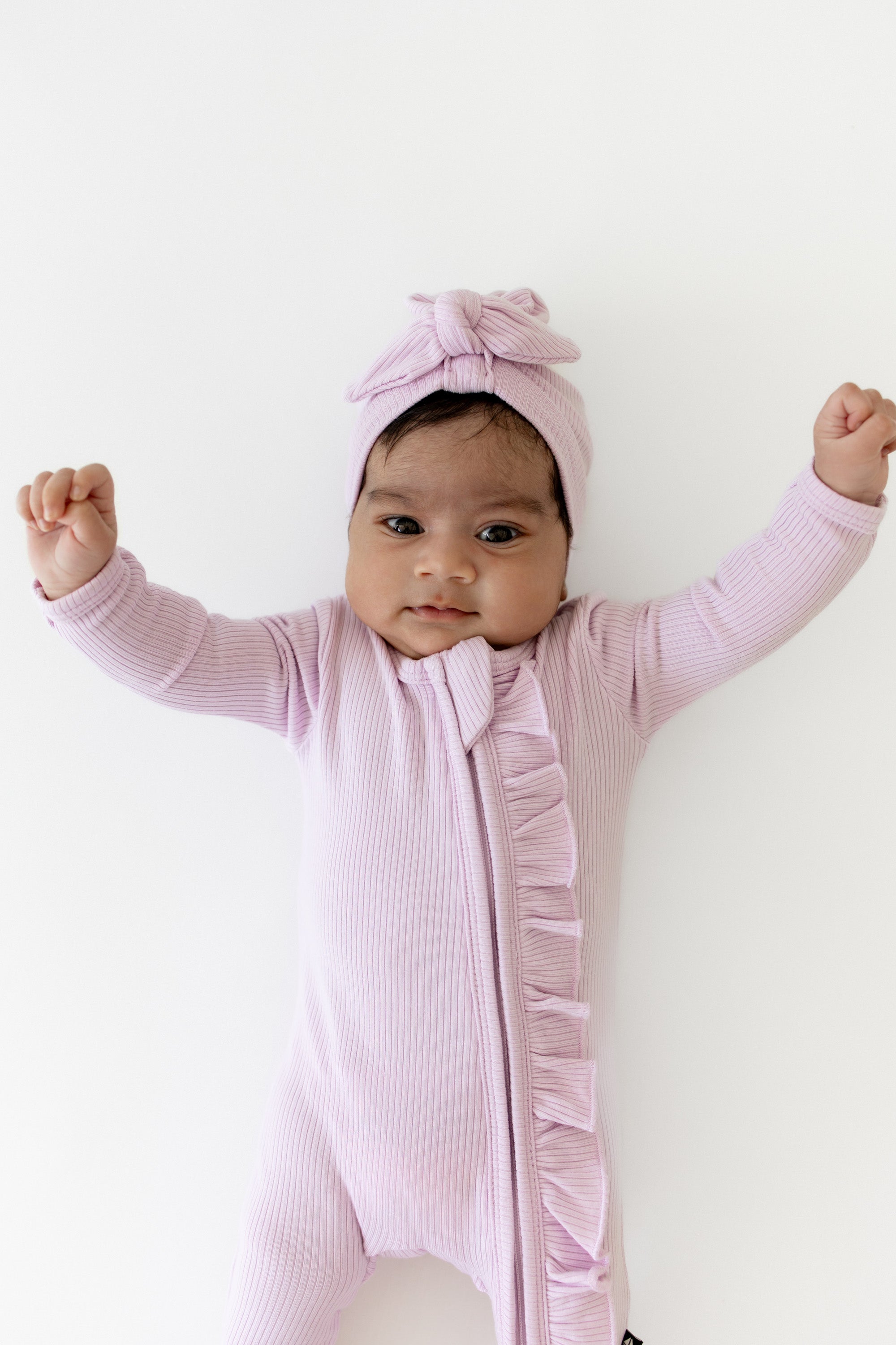Young infant stretching wearing the Ribbed Ruffle Zipper Footie in Thistle paired with a matching ribbed headwrap