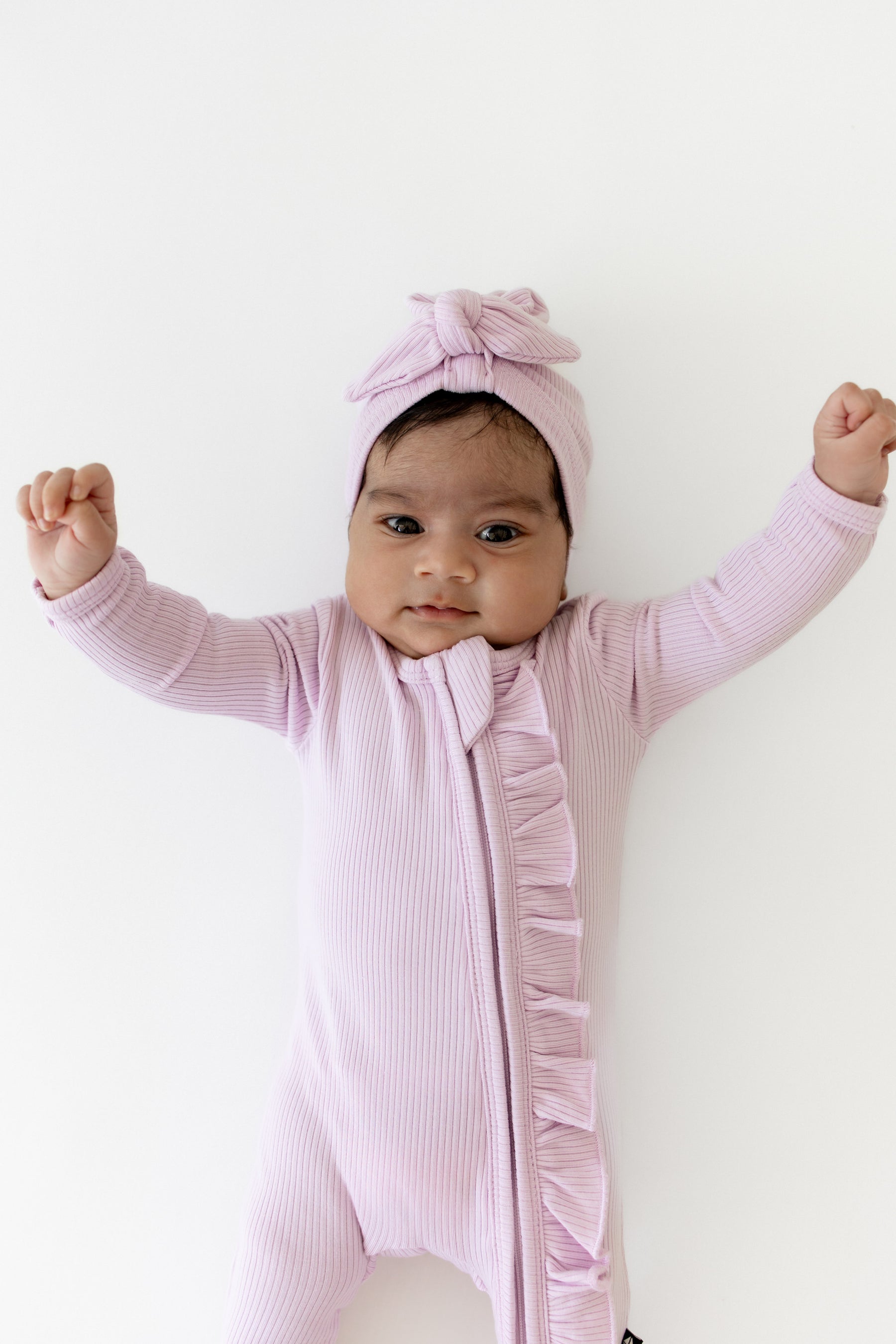 Young infant stretching wearing the Ribbed Ruffle Zipper Footie in Thistle paired with a matching ribbed headwrap
