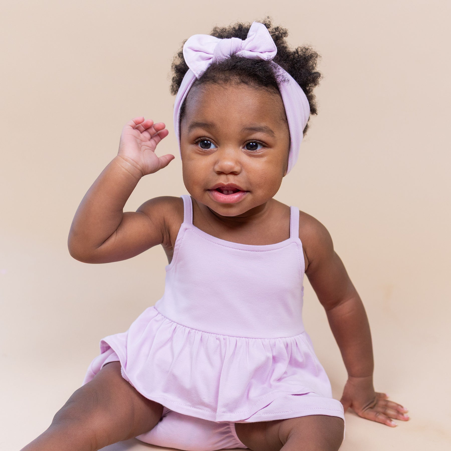 Young toddler sitting on the floor wearing the Spaghetti Strap Ruffle Romper in Thistle with matching bow