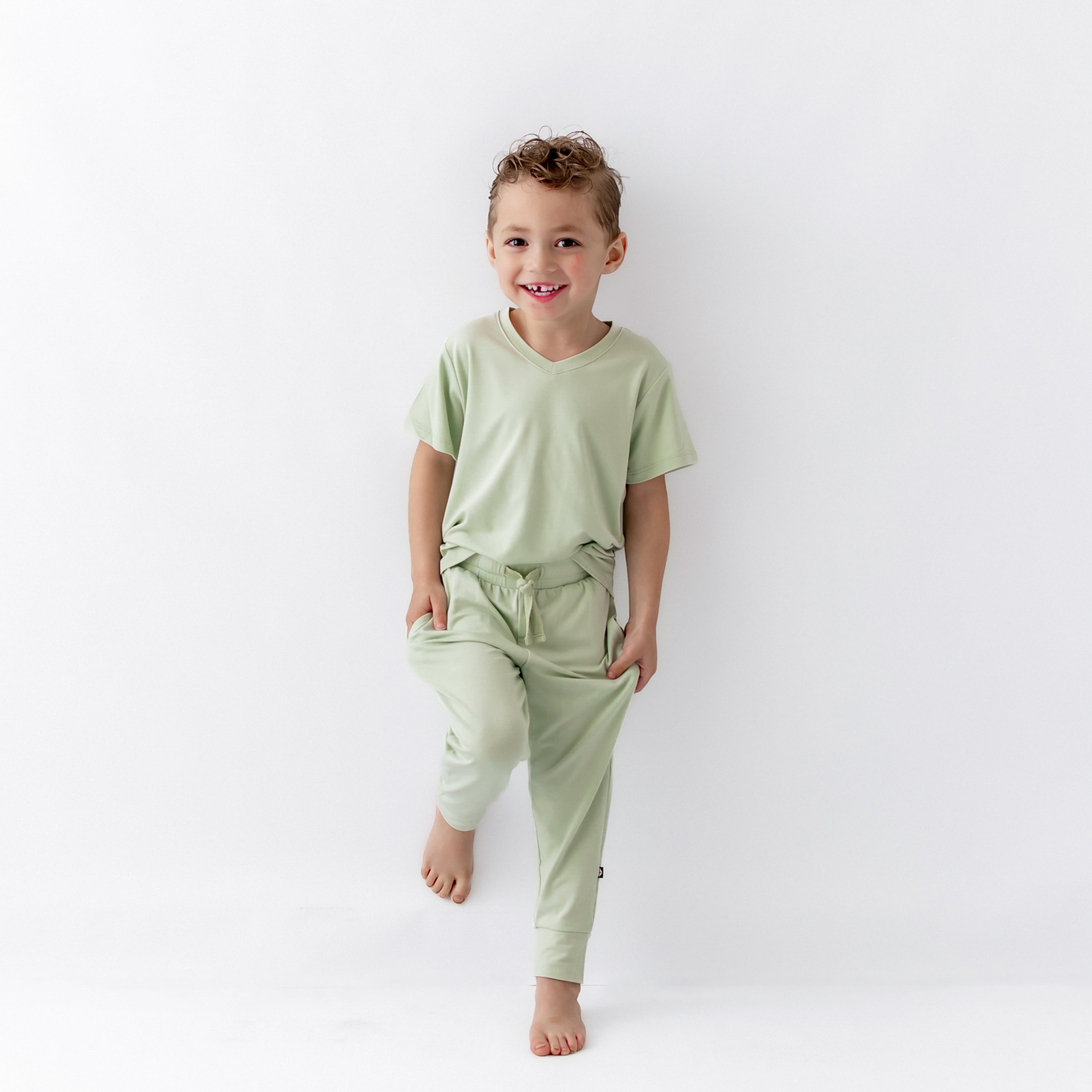Young boy leaning against a wall wearing the Toddler Jogger Pant in Basil paired with matching toddler V-Neck