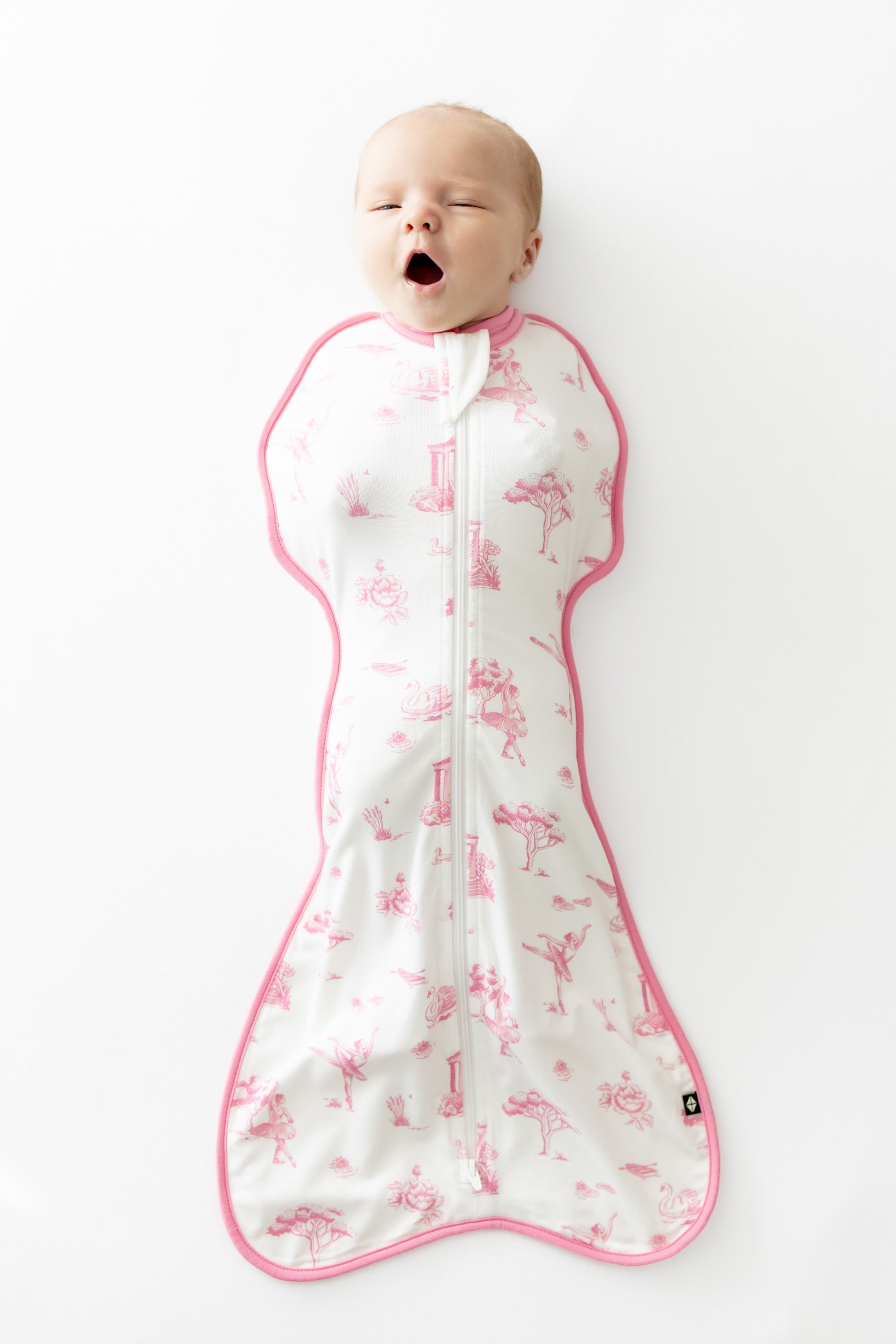 Yawning baby wearing the Swaddle Bag in Toile Ballet 0.5 laying on a white background
