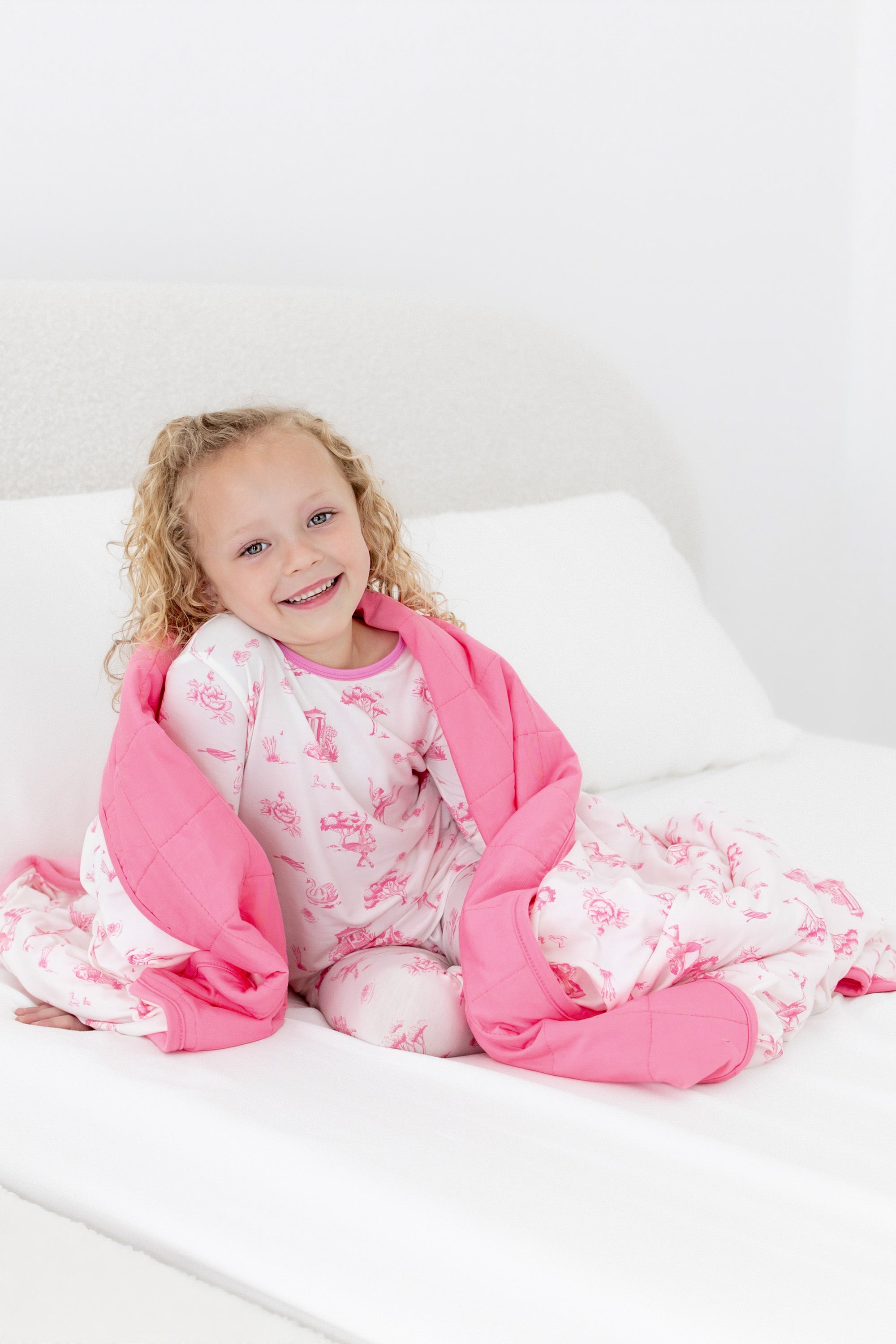 Young girl sitting on a bed with the Toddler Blanket in Toile Ballet 1.0 wrapped around her shoulder wearing matching long sleeve pajamas