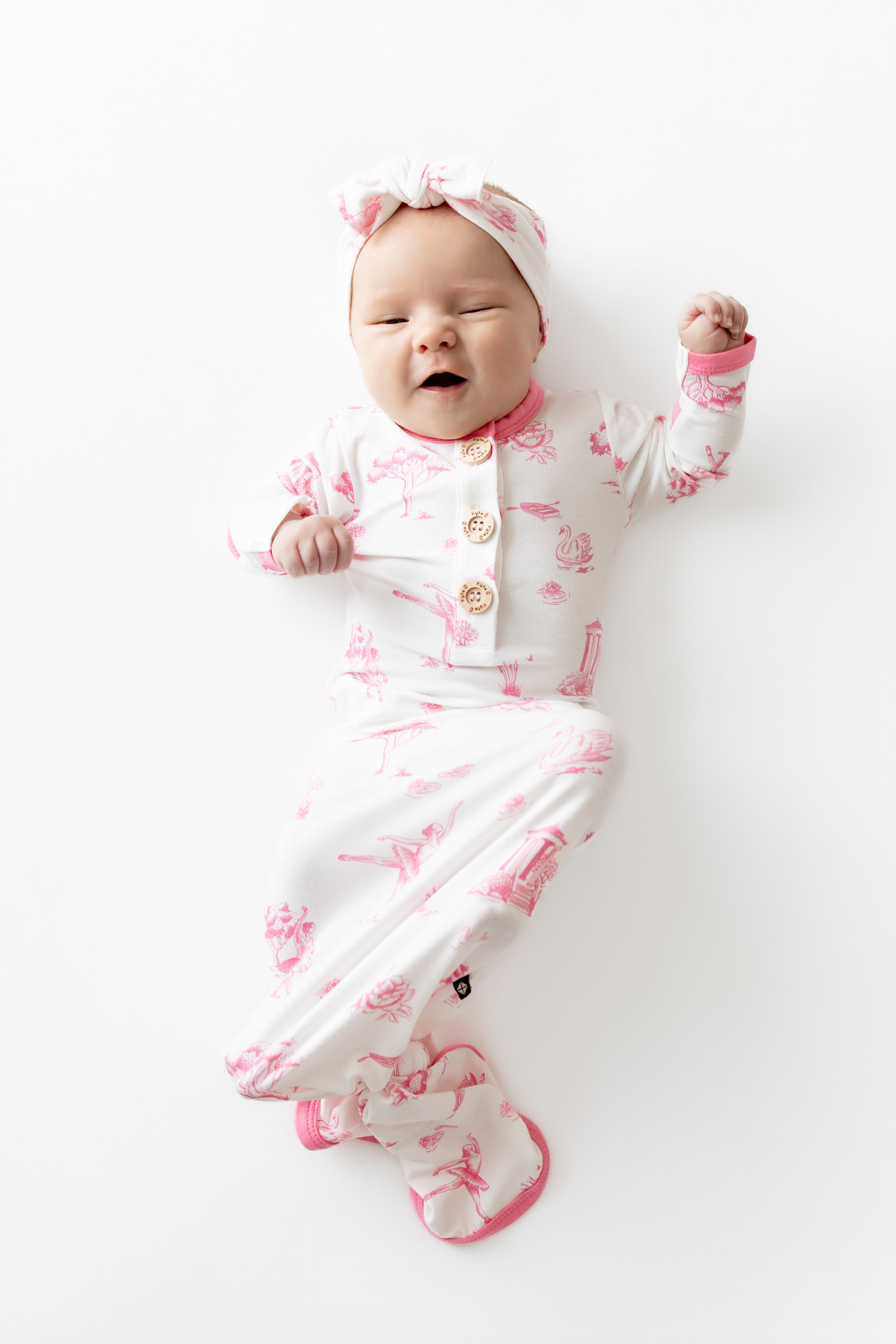 Infant girl laying on a white surface wearing the Knotted Gown with Bow Set in Toile Ballet