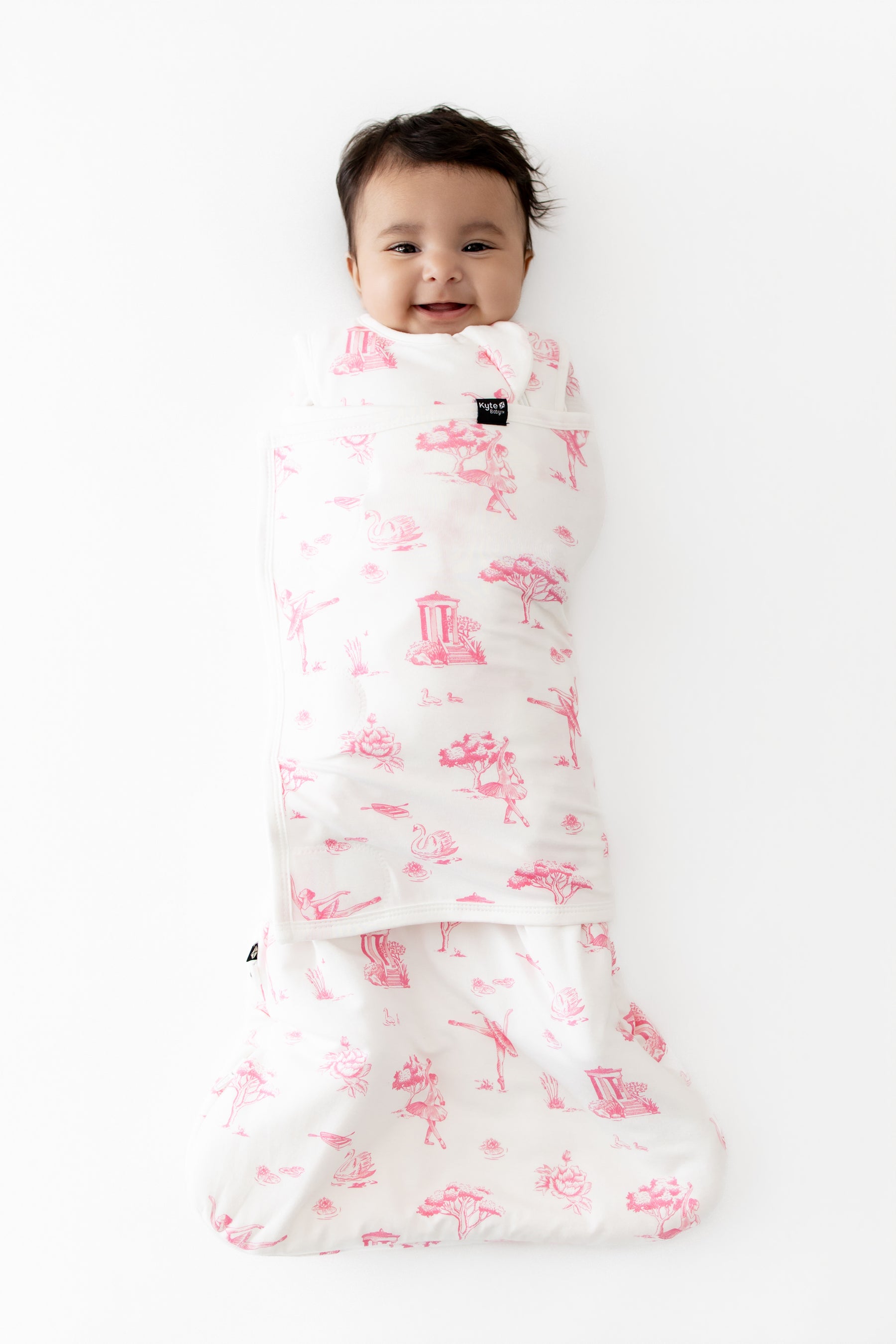 Young infant laying on a white surface swaddled in the Sleep Bag Swaddler in Toile Ballet 1.0