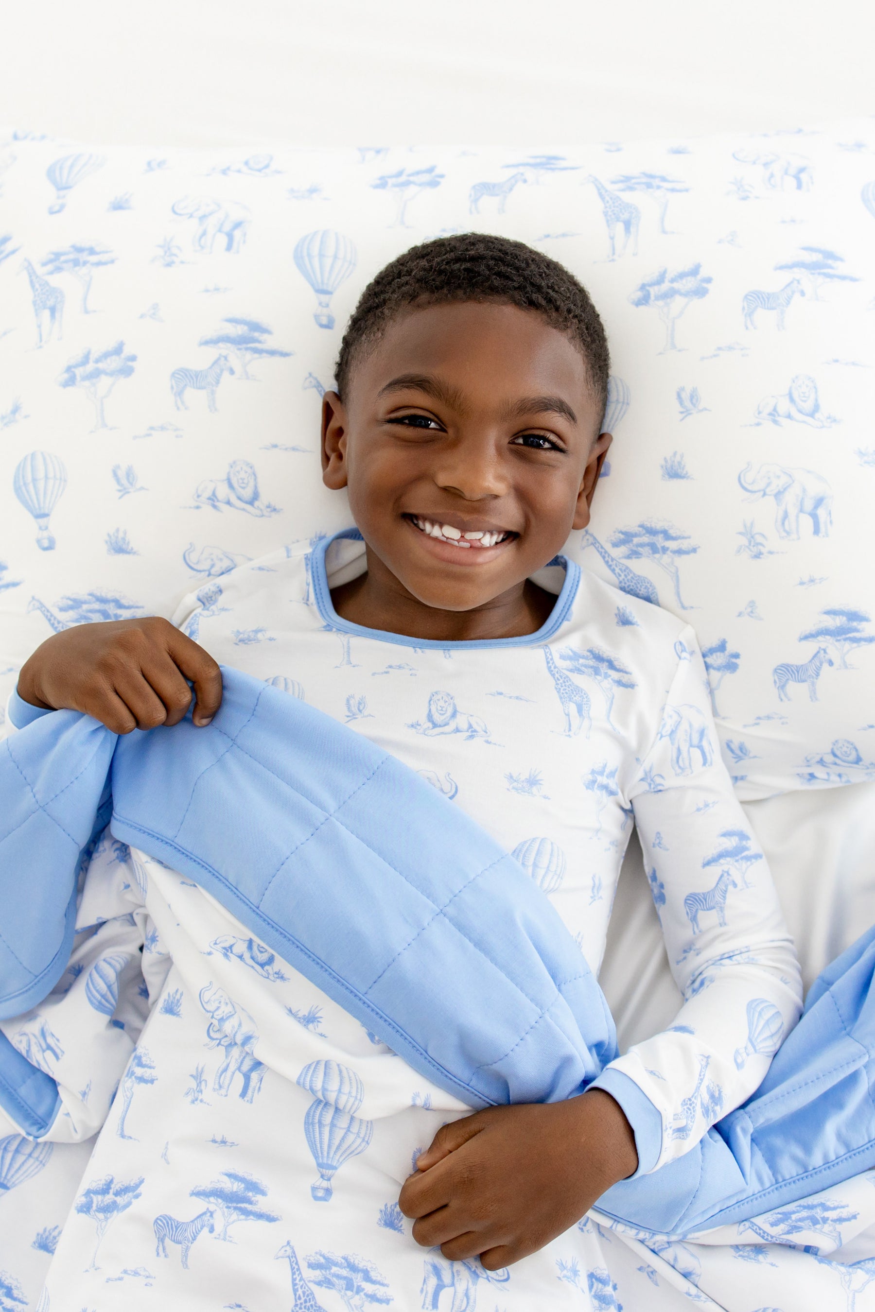 Young boy laying in bed with the Toddler Blanket in Toile Safari 1.0 overtop of him laying on a pillow with the Standard Pillow case in Toile Safari