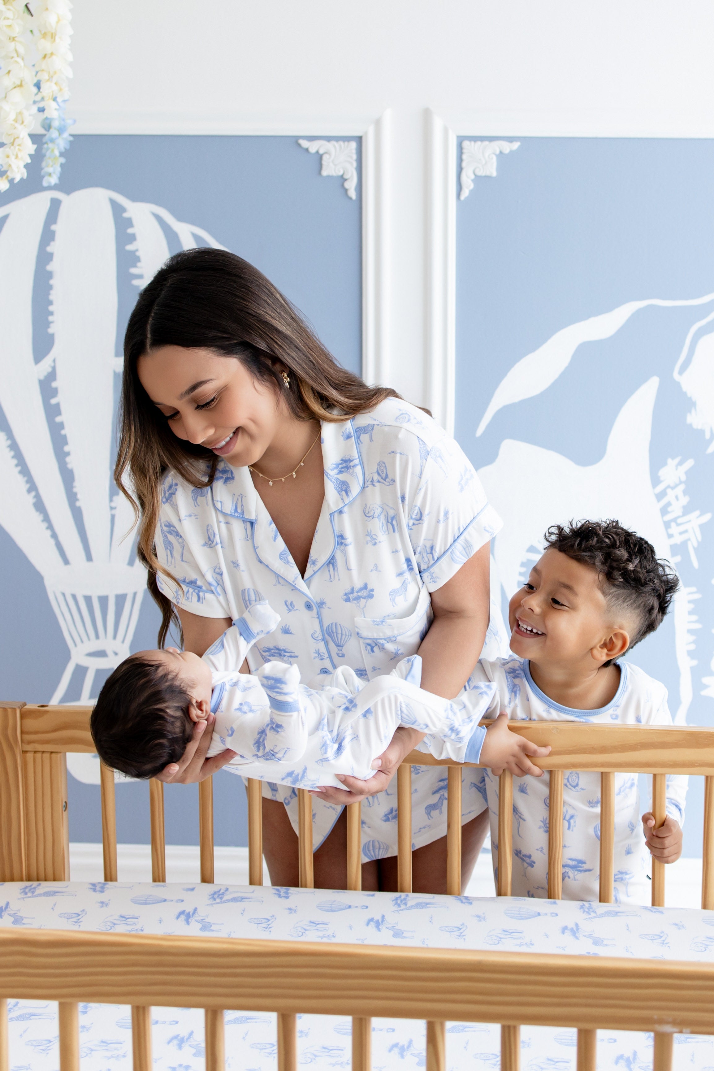 Woman holding a baby in front of a smiling child in a crib with a blue wall featuring hot air balloon artwork.