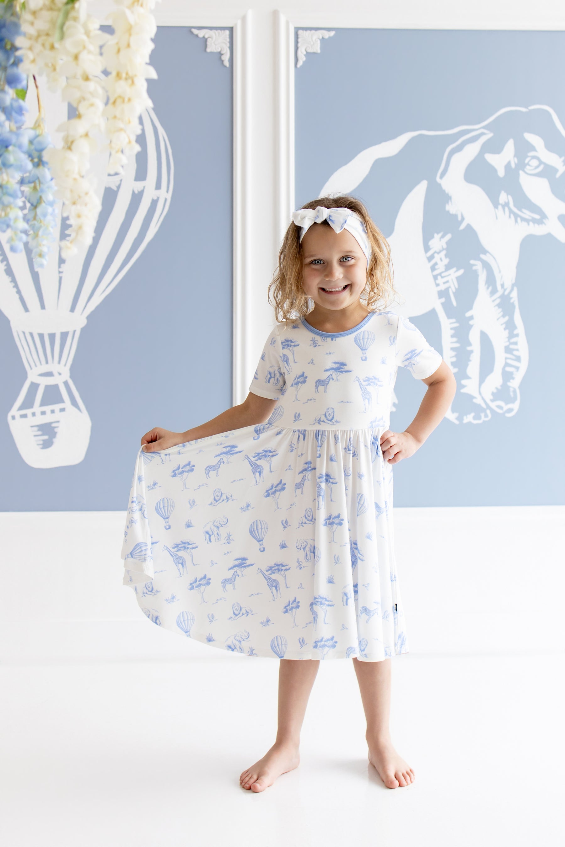Young girl modeling the Twirl Dress in Toile Safari holding the skirt of the dress out standing in front of safari inspired artwork