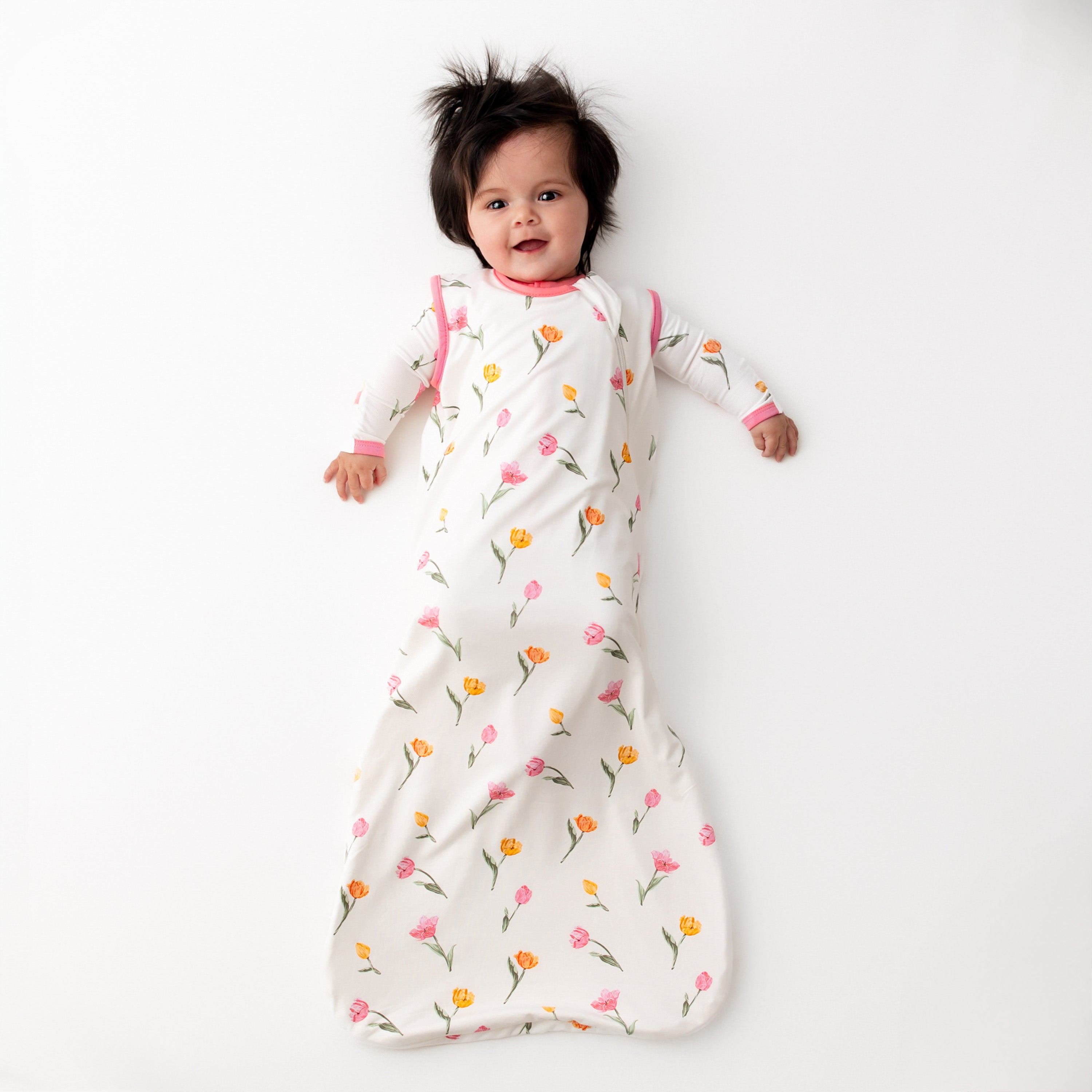 Smiling toddler laying down wearing the Sleep Bag in Tulip 0.5 with a matching long sleeve romper on underneath