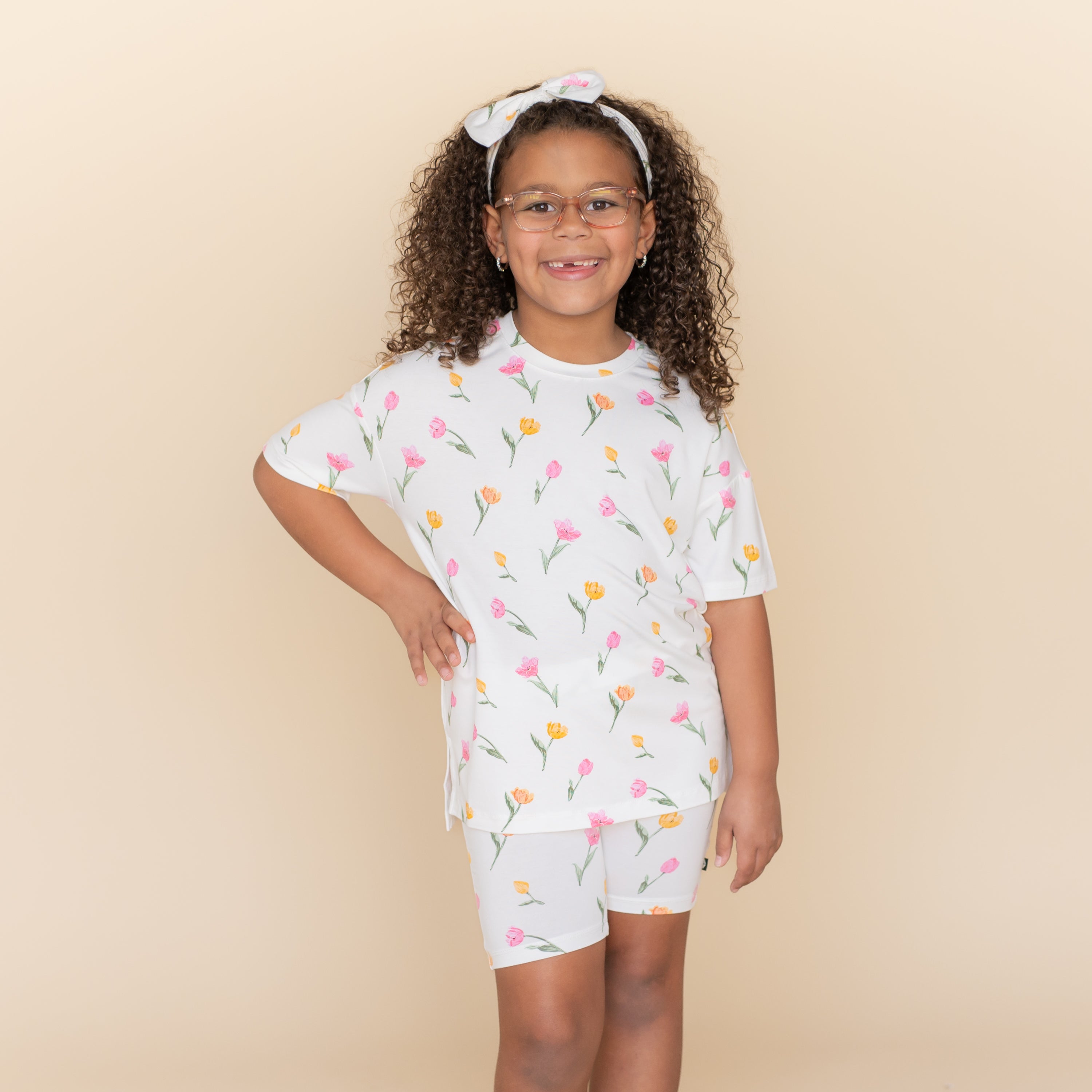 Young girl posing wearing the Toddler Biker Short Set in Tulip with a matching bow standing in front of a light taupe background