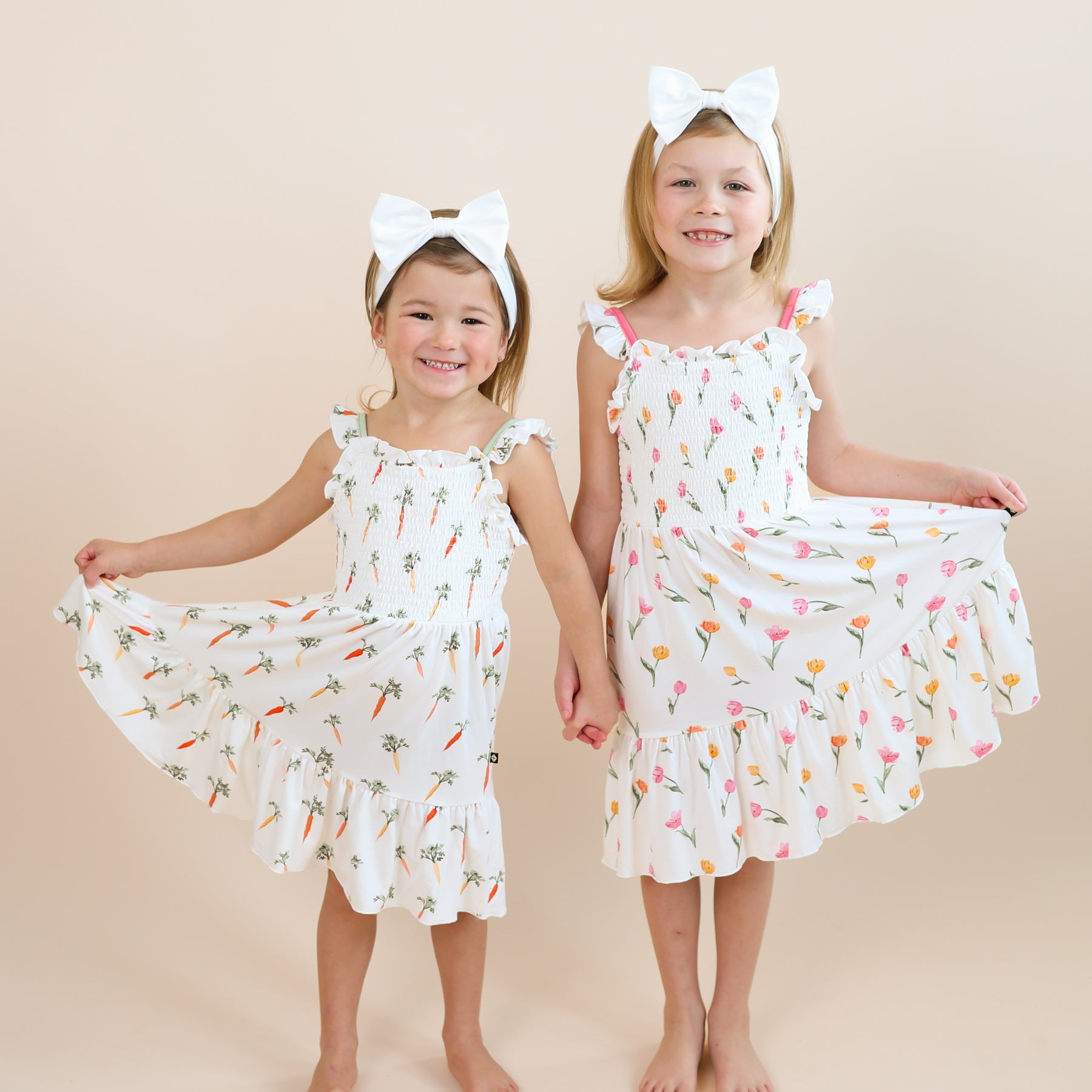 Two sisters standing ide by side holding hands wearing the Smocked Dress in Tulip and Carrot