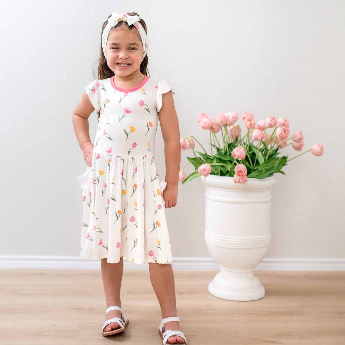 Young girl posing wearing the Pocket Dress in Tulip standing beside a white vase with pink flowers