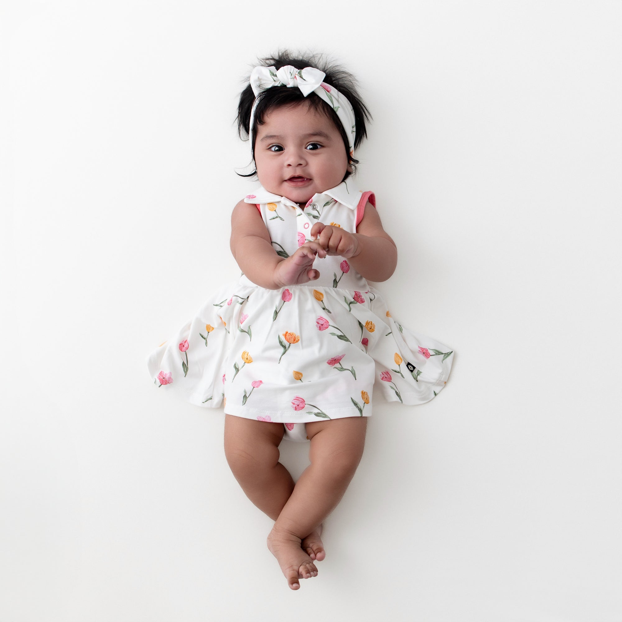 Infant laying on a white surface wearing the Polo Bodysuit Dress in Tulip with matching knotted bow headband