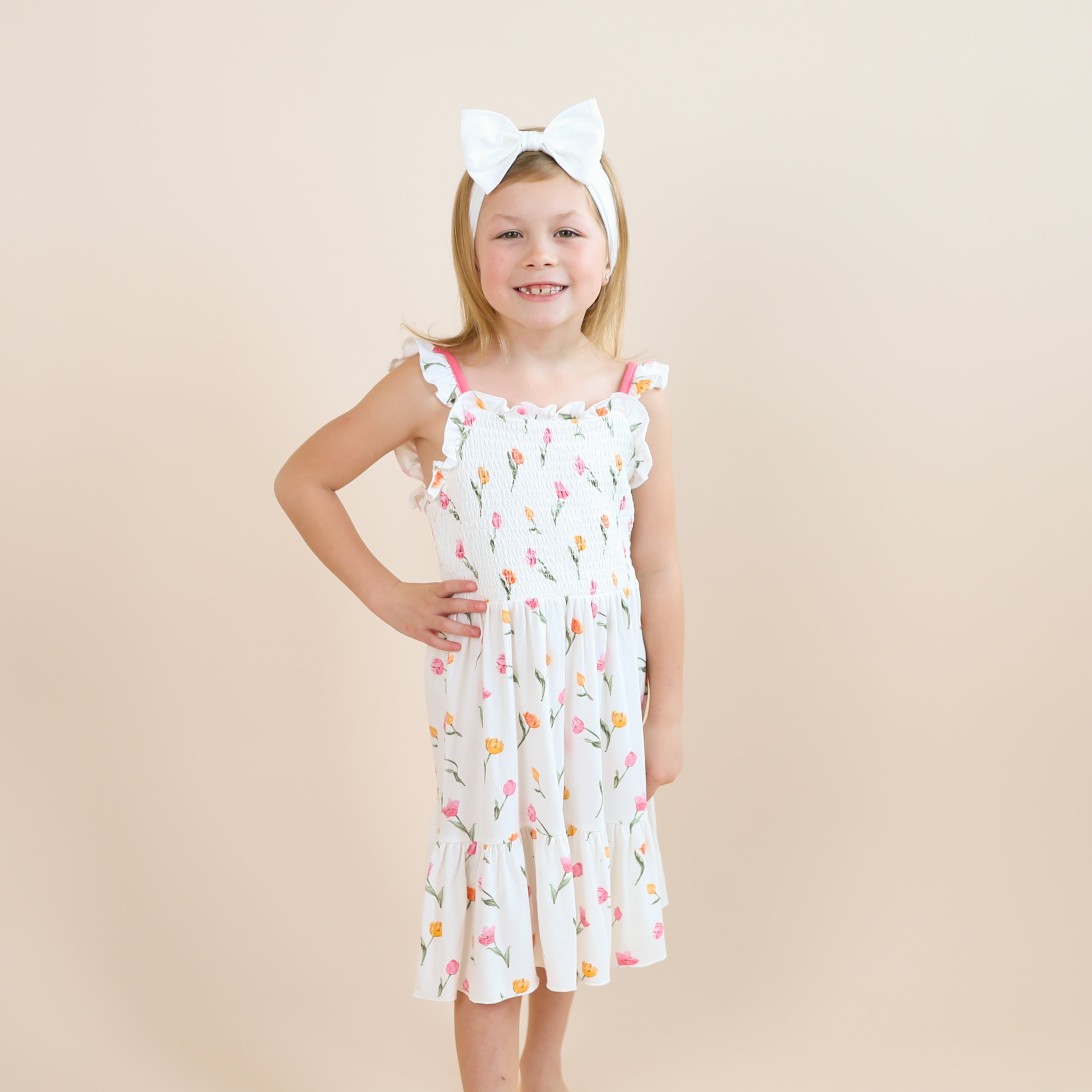 Smiling girl posing wearing the Smocked Dress in Tulip paired with a white bow standing in front of a light taupe background