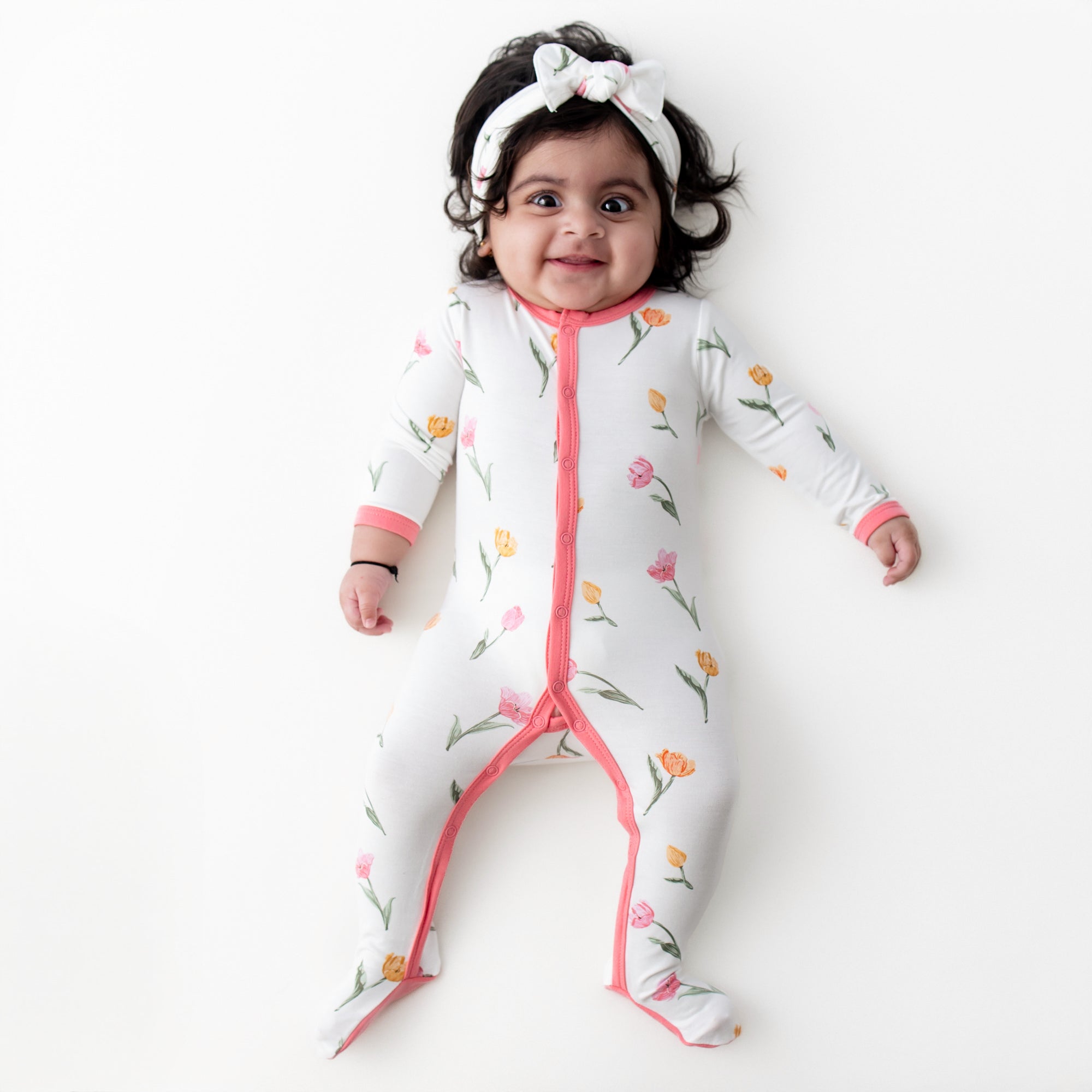 Young infant laying on a white surface wearing the Footie in Tulip with matching knotted bow headband