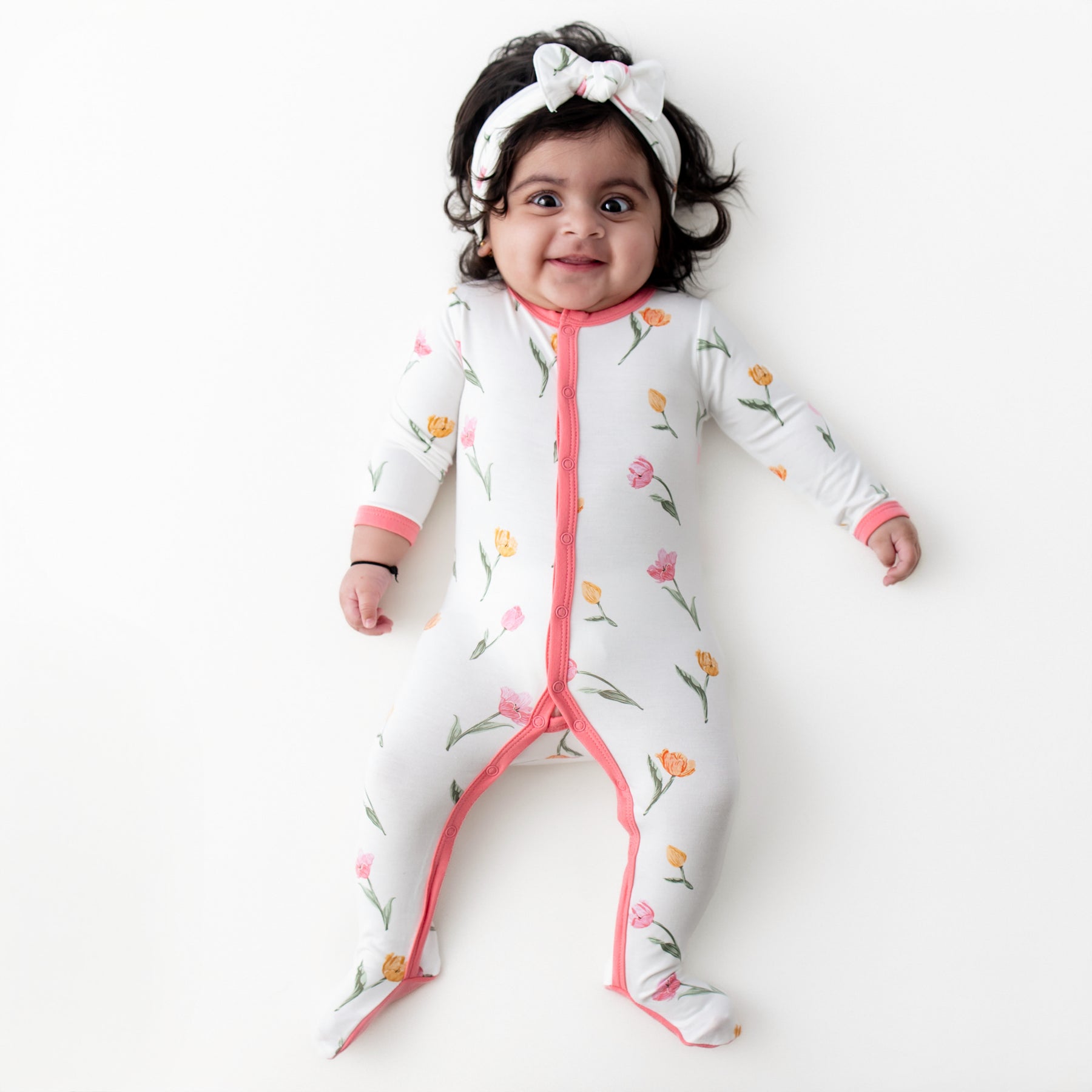Young infant laying on a white surface wearing the Footie in Tulip with matching knotted bow headband