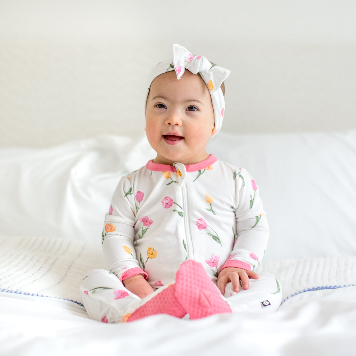 Young toddler sitting on a bed wearing the Zippered Footie in Tulip showing the pink colored foot pads paired with a matching bow