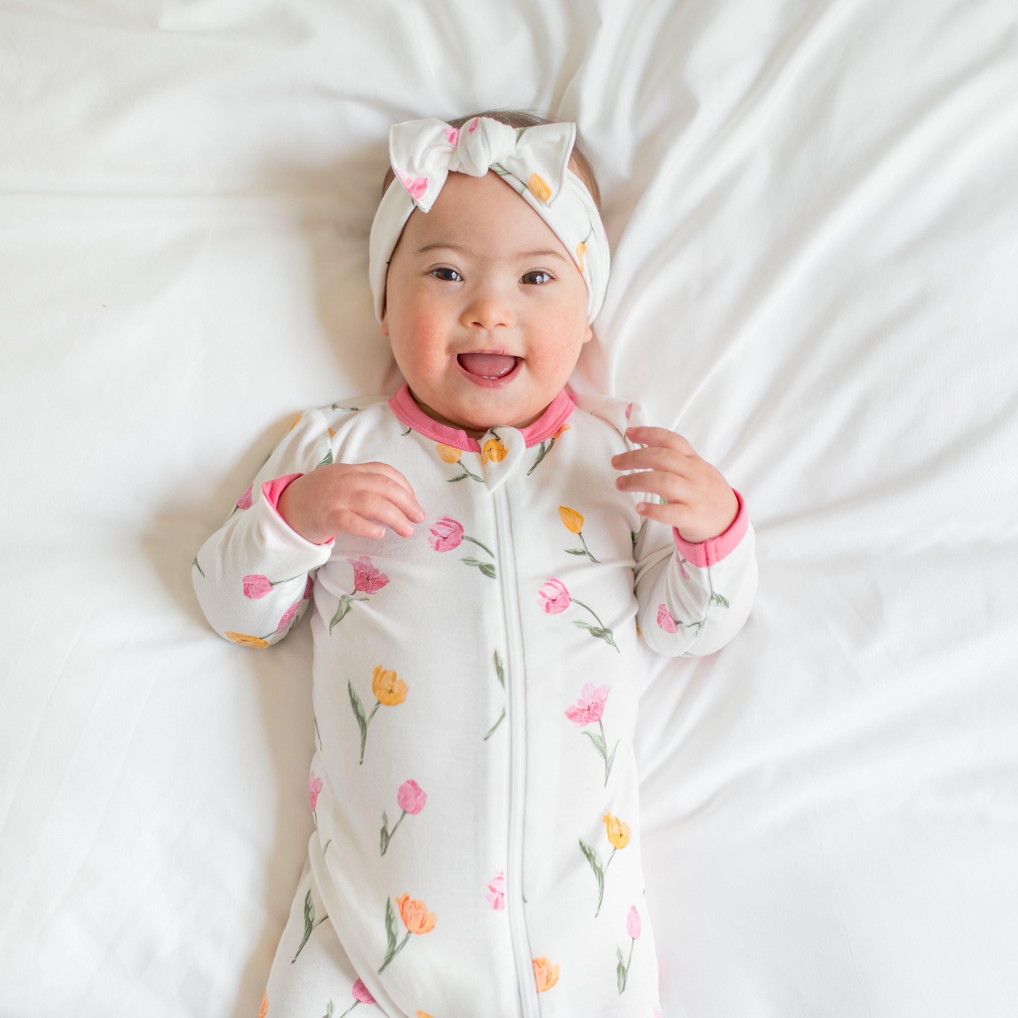 Close up of smiling toddler wearing the Zippered Footie in Tulip and matching bow laying on a bed