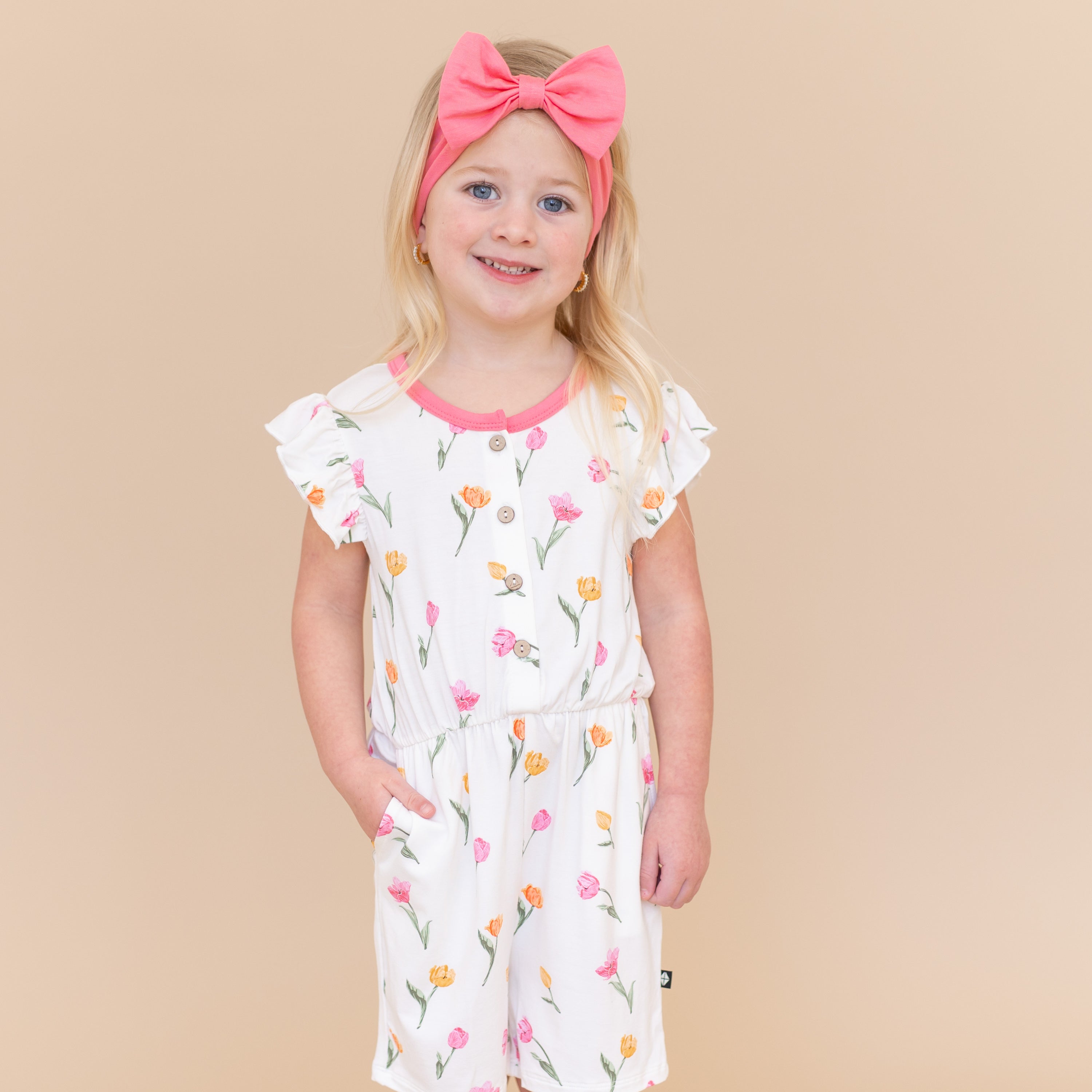 Young girl posing wearing the Flutter Sleeve Romper in Tulip paired with a pink bow