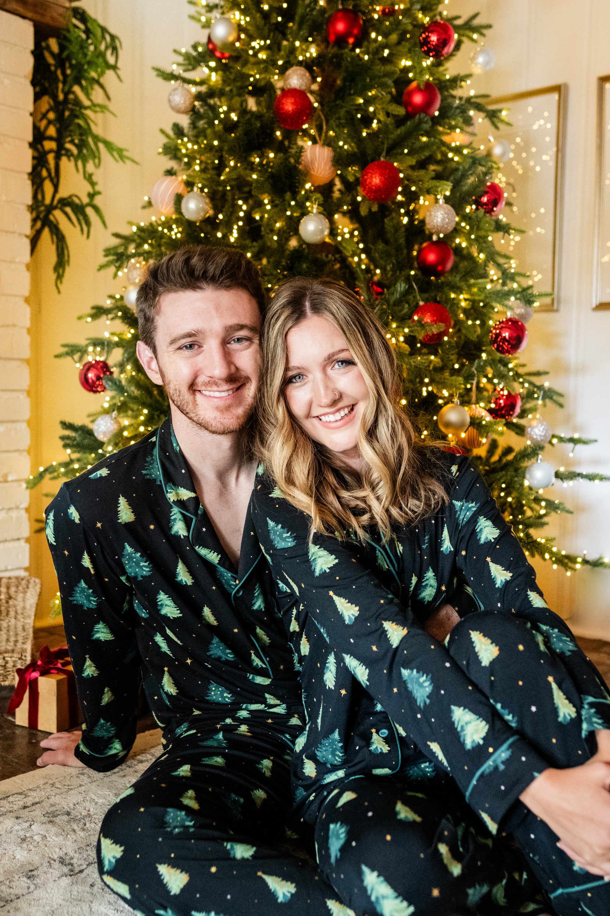 Husband and wife sitting on the floor in front of a Christmas tree both matching in Twinkle Tree Pajama Sets