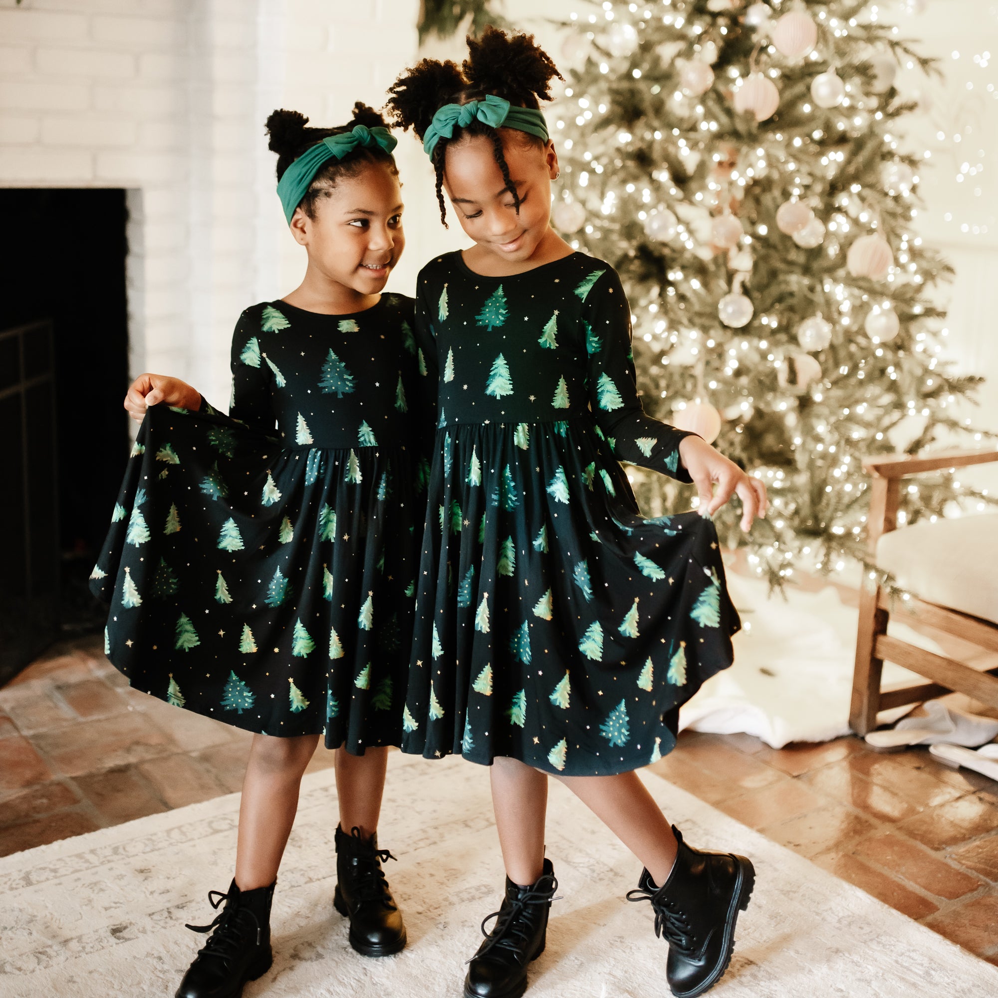 Two sisters standing side by side holding the hems of their Long Sleeve Twirl Dress in Twinkle Tree in front of a decorated Christmas tree
