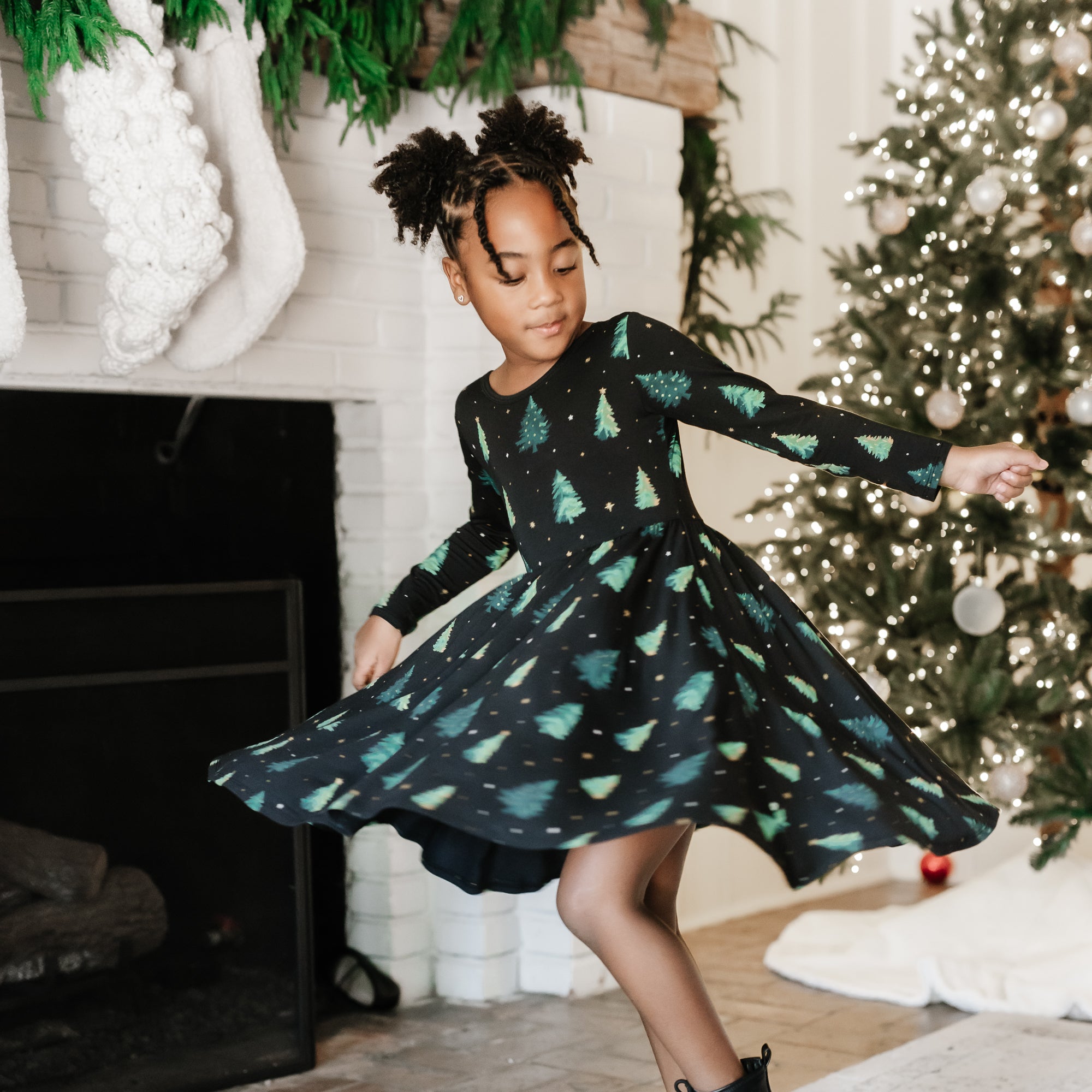 Young girl twirling in the Long Sleeve Twirl Dress in Twinkle Tree standing beside a white fireplace in front of a decorated Christmas tree