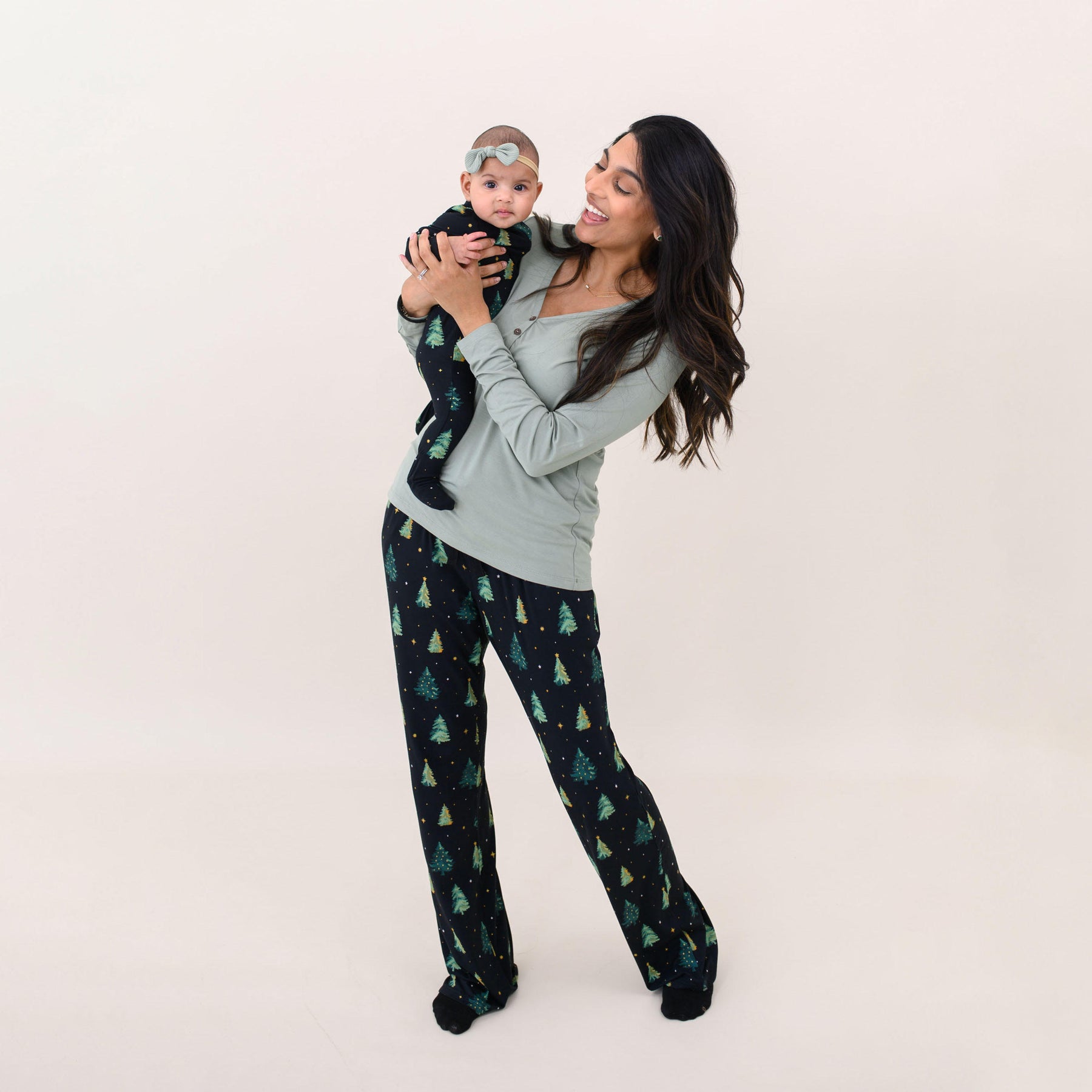 Mother wearing the Women's Twinkle Tree Lounge pants and Thyme Henley shirt holding her daughter wearing the Zippered Footie in Twinkle Tree