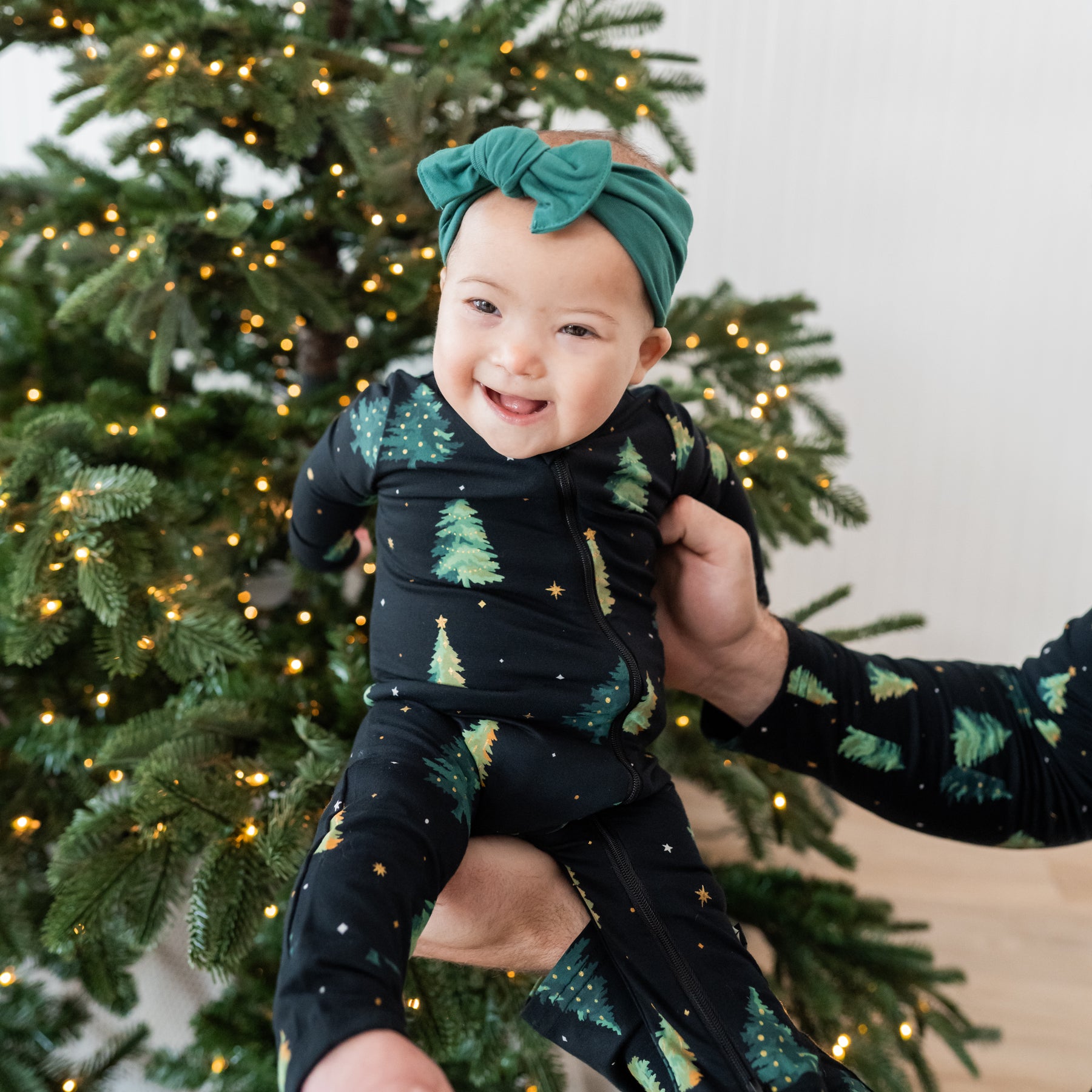 Smiling infant girl being held by her father in front of a christmas tree wearing the Zippered Romper in Twinkle Tree and Emerald knotted bow headband